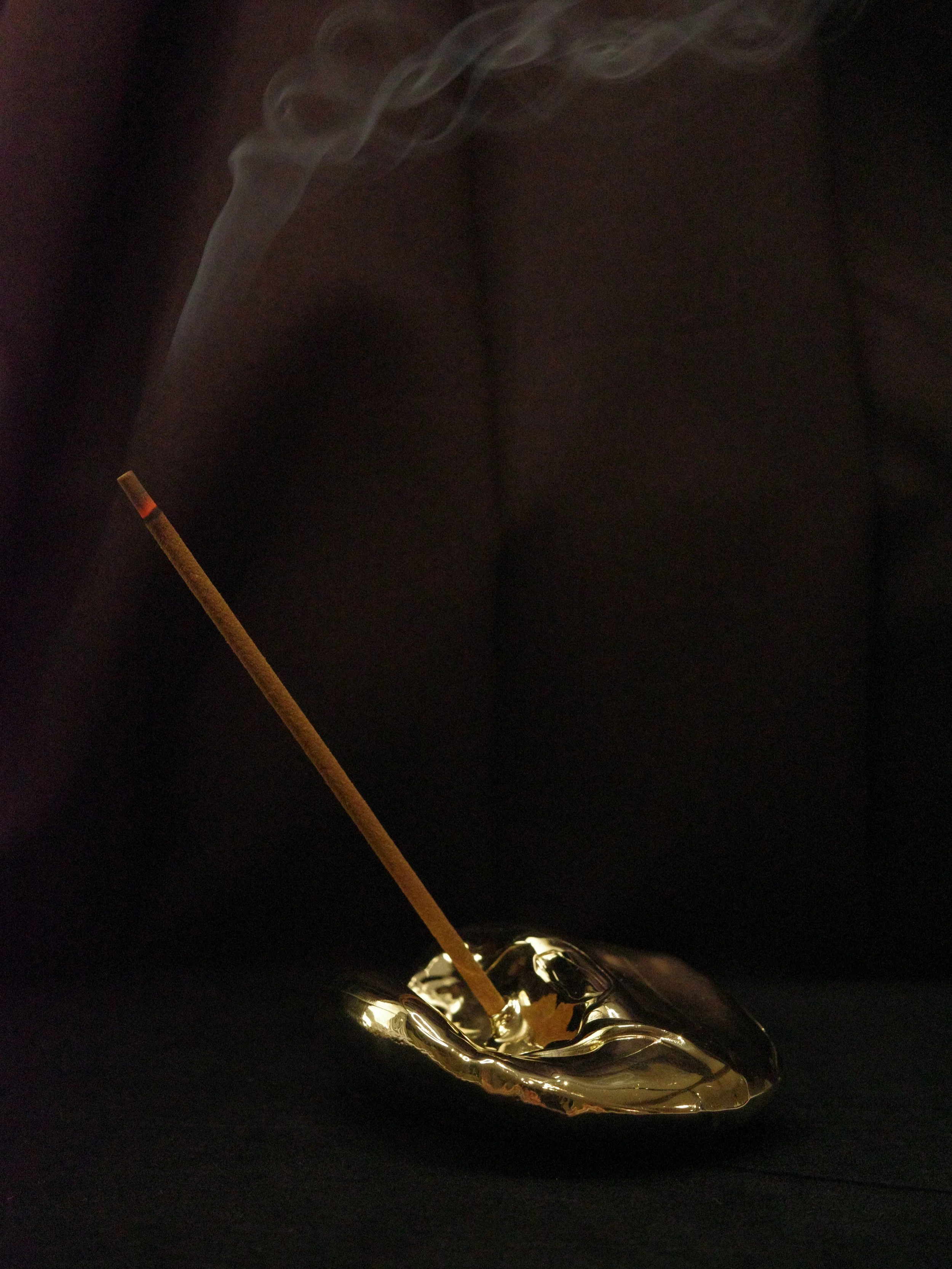 An unlit incense stick placed in a gold-colored ceramic ashtray with smoke rising from the burning tip.