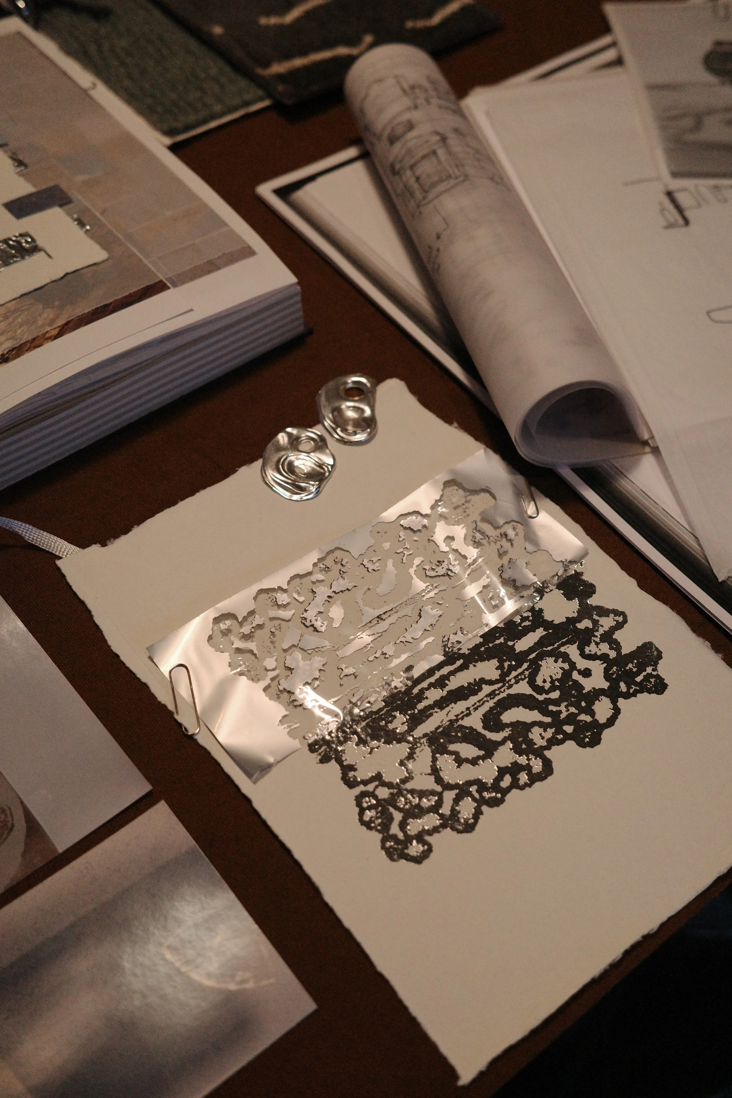 A table with a foil stencil, two shiny metal embellishments, and open design books, likely related to art or crafts.