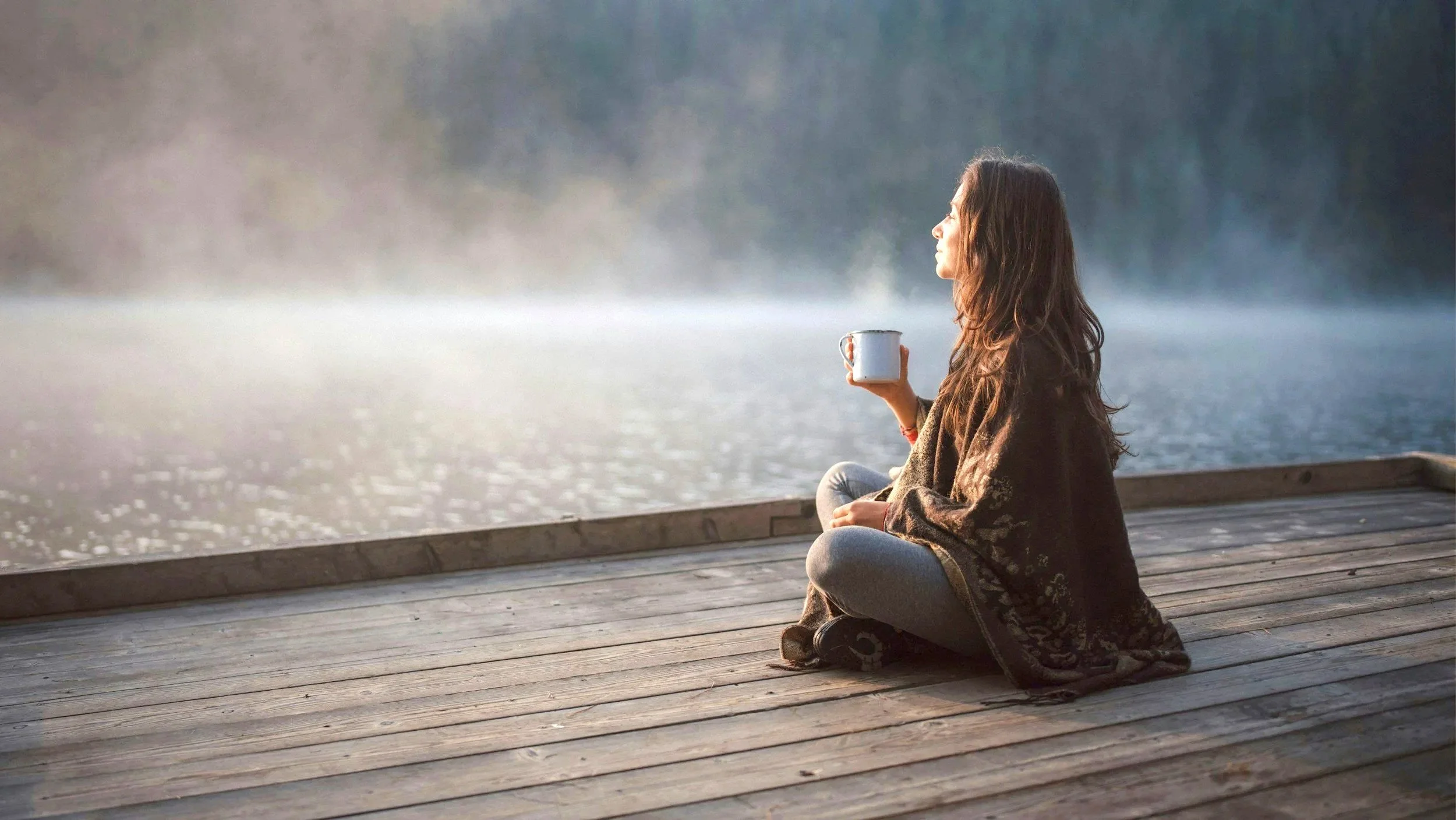 Woman sitting peacefully beside a lake, holding a mug and looking out across the water