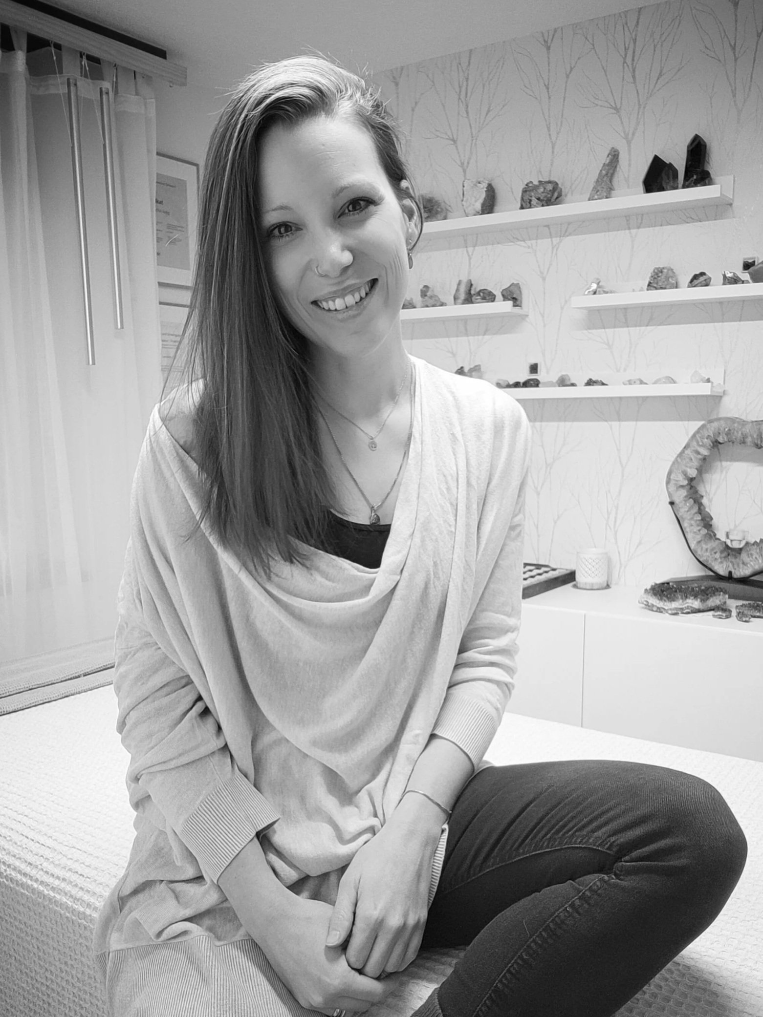 Schwarz-Weiß-Foto einer lächelnden Frau mit langem Haar, bequem gekleidet, sitzend auf einem Bett in einem modernen Zimmer mit dekorativen Felsen und Mineralien auf Regalen im Hintergrund.