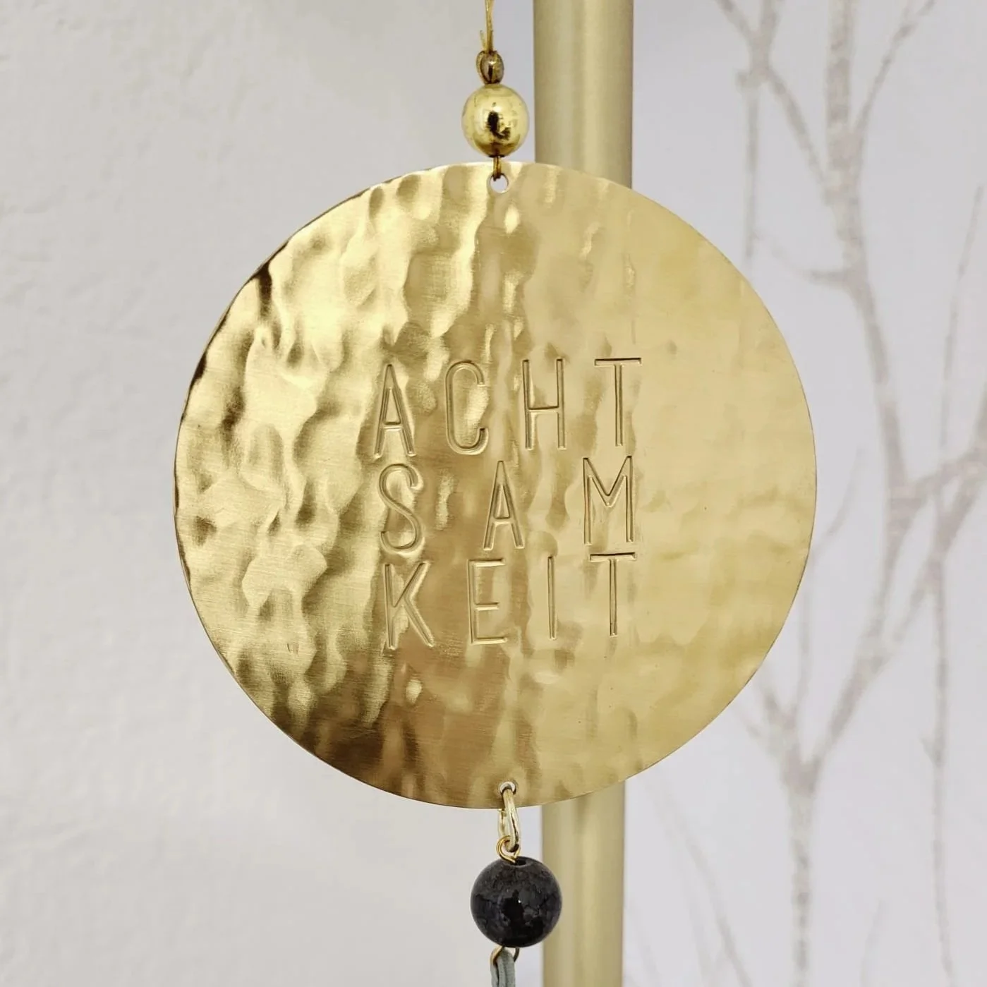 Goldene Windspiel mit eingraviertem Text »ACHT SAM KET«, hängt an einer Kette, mit einem schwarzen Kristall am unteren Ende.