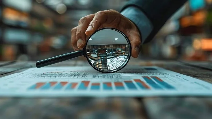 Person holding a magnifying glass over printed charts and graphs on a table in a bookstore or library.