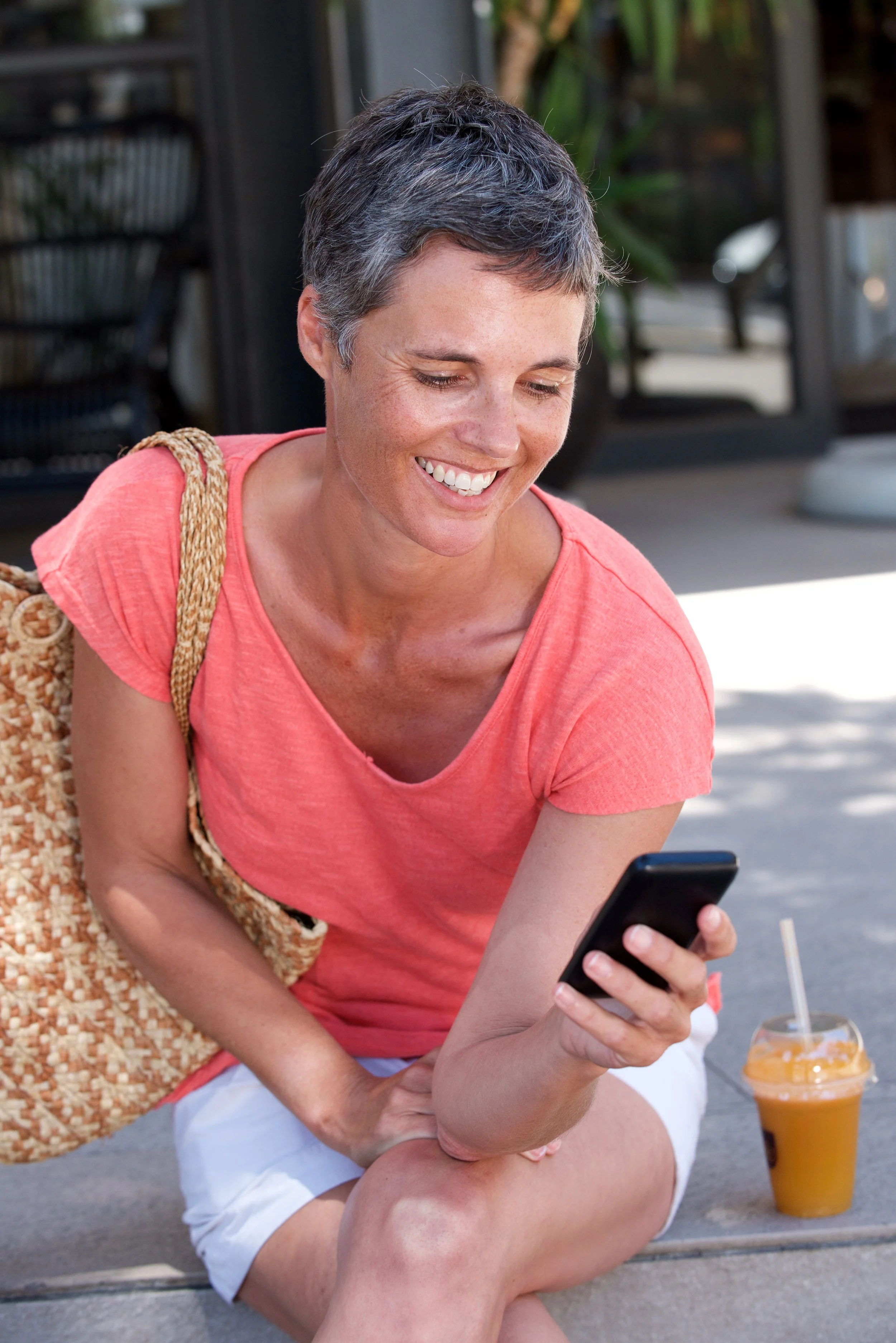 Una mujer con cabello corto y tez clara, vestida con camiseta rosa y shorts blancos, está sentada en la acera mirando y sonriendo a su teléfono móvil, con una bebida de jugo de naranja al lado.