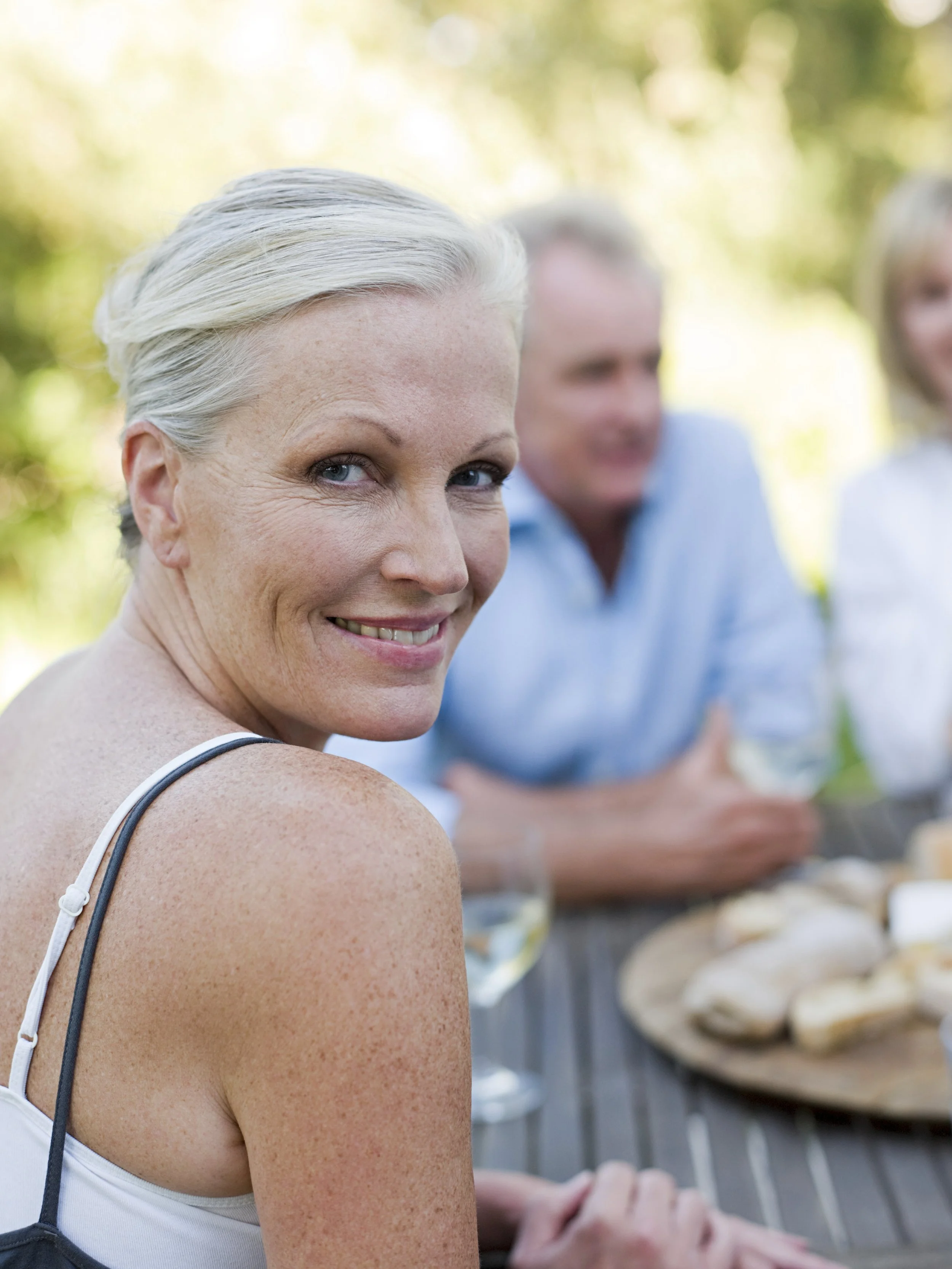 Mujer de cabello gris sonriendo en una reunión al aire libre con amigos, en un día soleado, en un entorno natural.