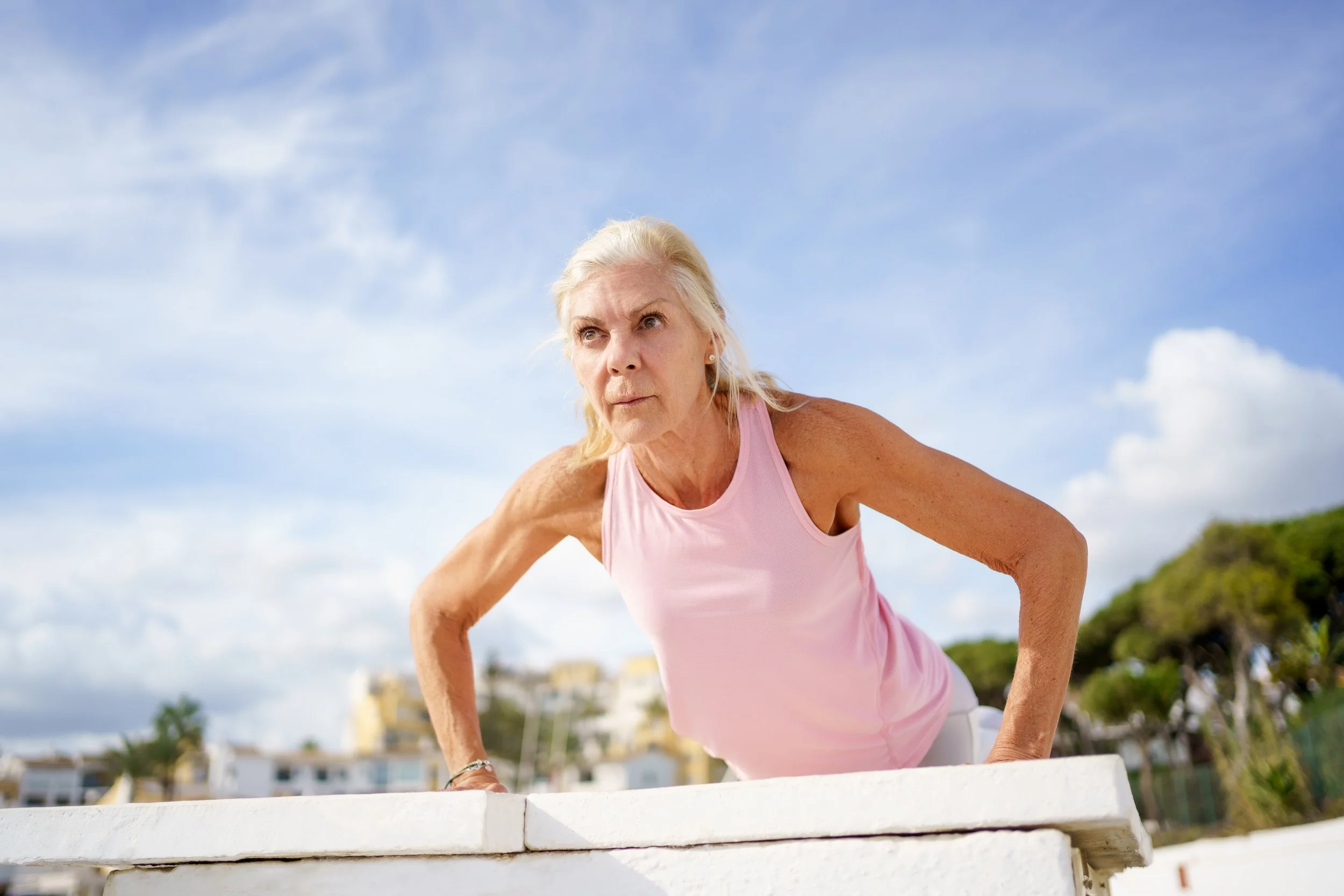 Mujer menopáusica haciendo ejercicio al aire libre, apoyada en una estructura blanca, con cielo y árboles al fondo.