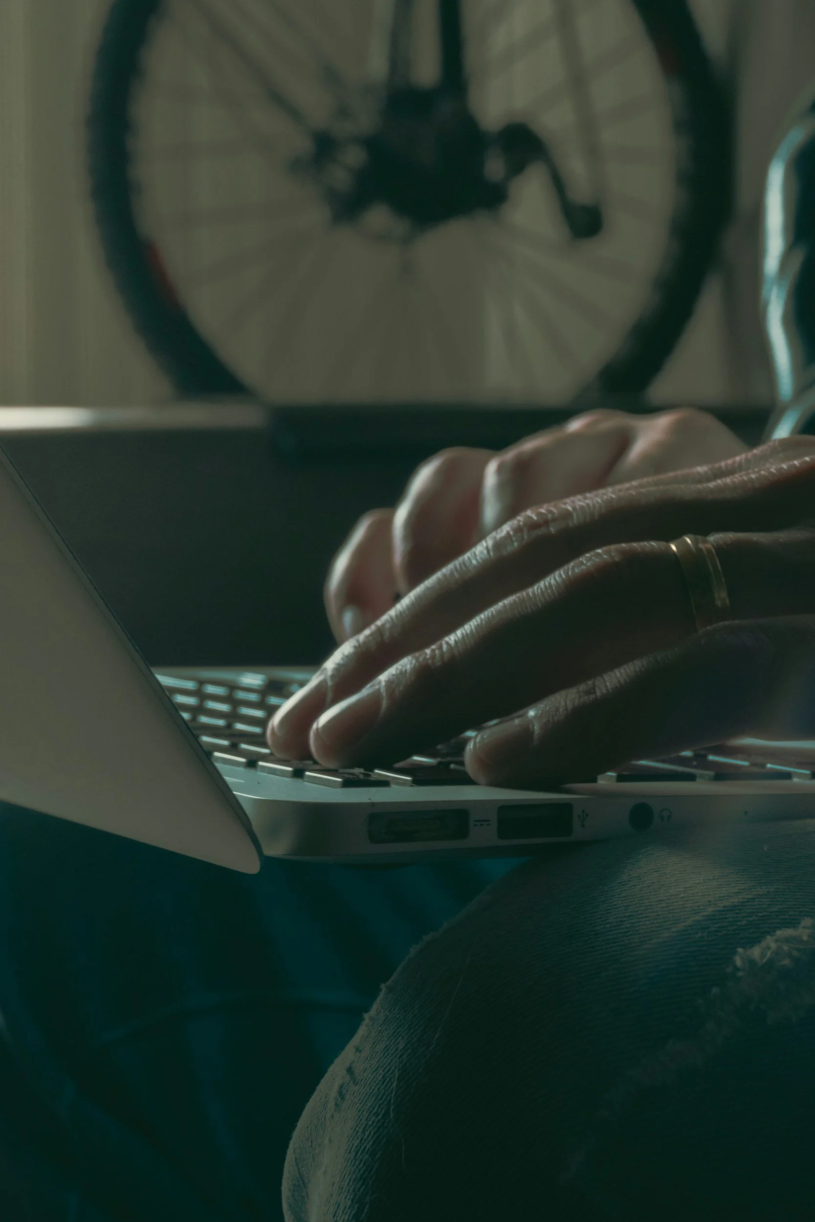 Close-up of a person's hand typing on a laptop keyboard with a bicycle rear wheel mounted on the wall in the background.