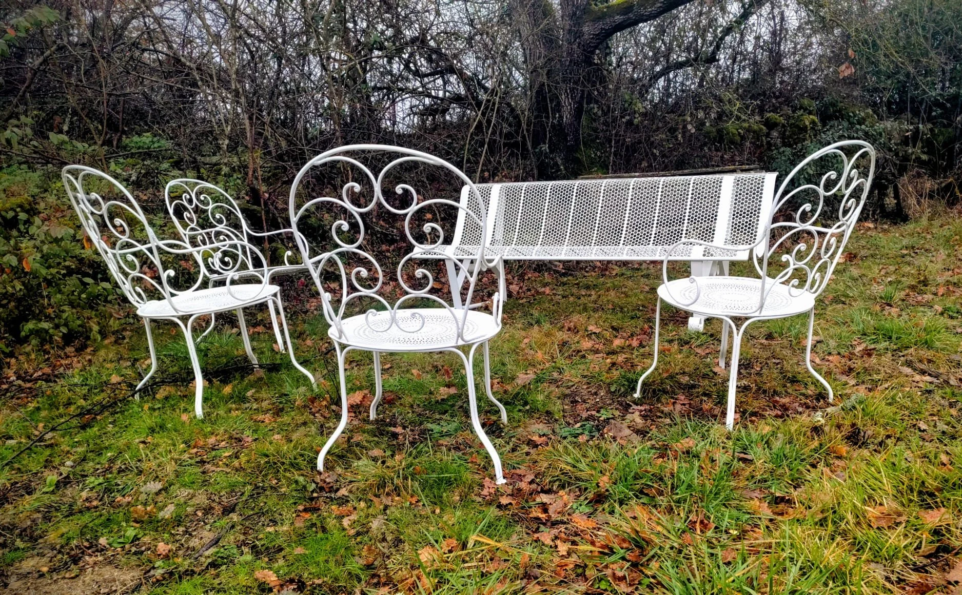 Ensemble de mobilier de jardin en fer blanc peint blanc, comprenant une table, une banquette et trois chaises avec des cadres en forme de volutes, situés dans un espace extérieur avec de l'herbe, des feuilles mortes, et des buissons en arrière-plan.