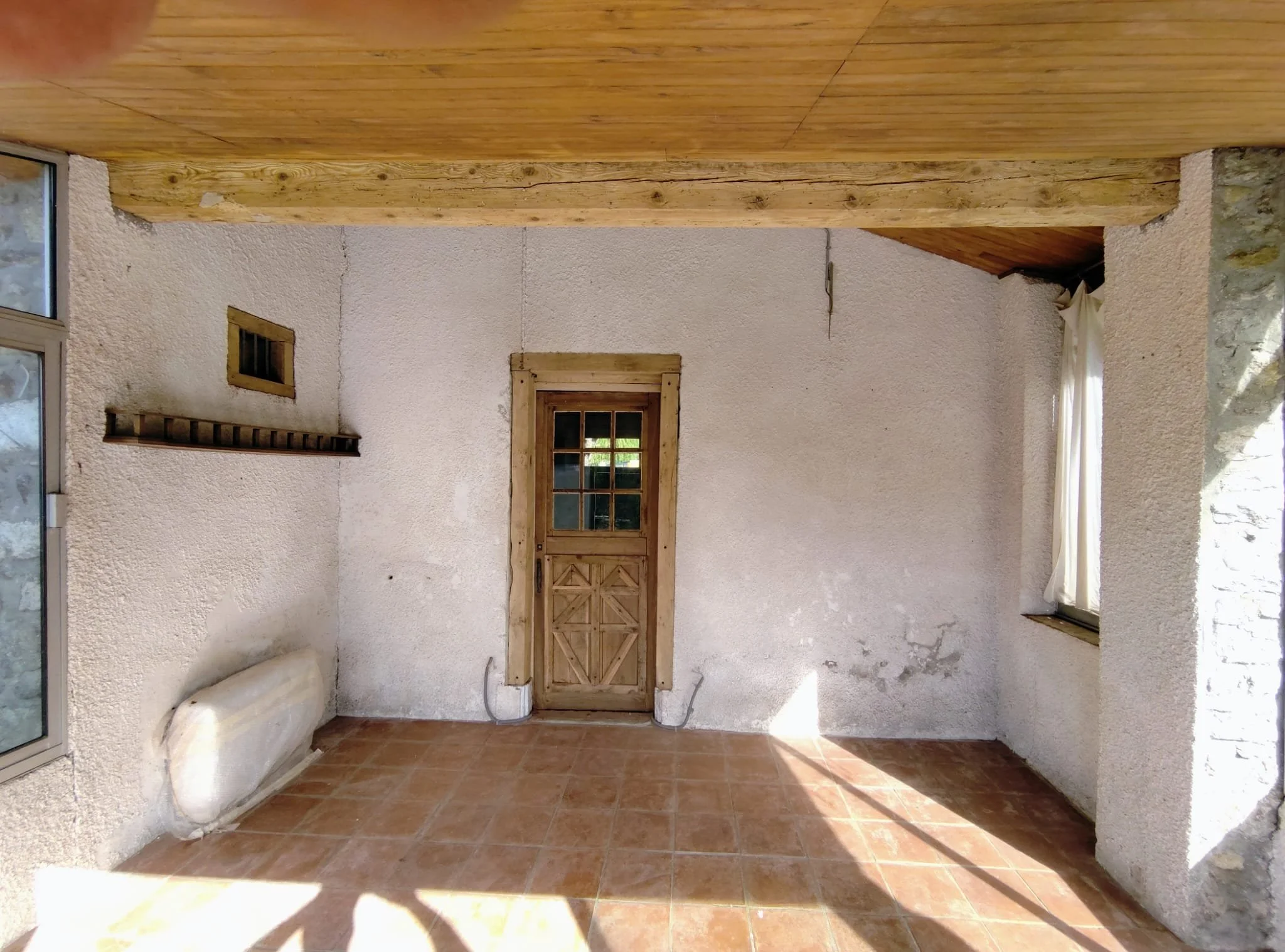 Pièce intérieure avec murs en plâtre blanc, porte en bois décapée au centre, une poutre décapée, une fenêtre en bois, plafond en lambris, sol en carreaux de terre cuite, et un radiateur .