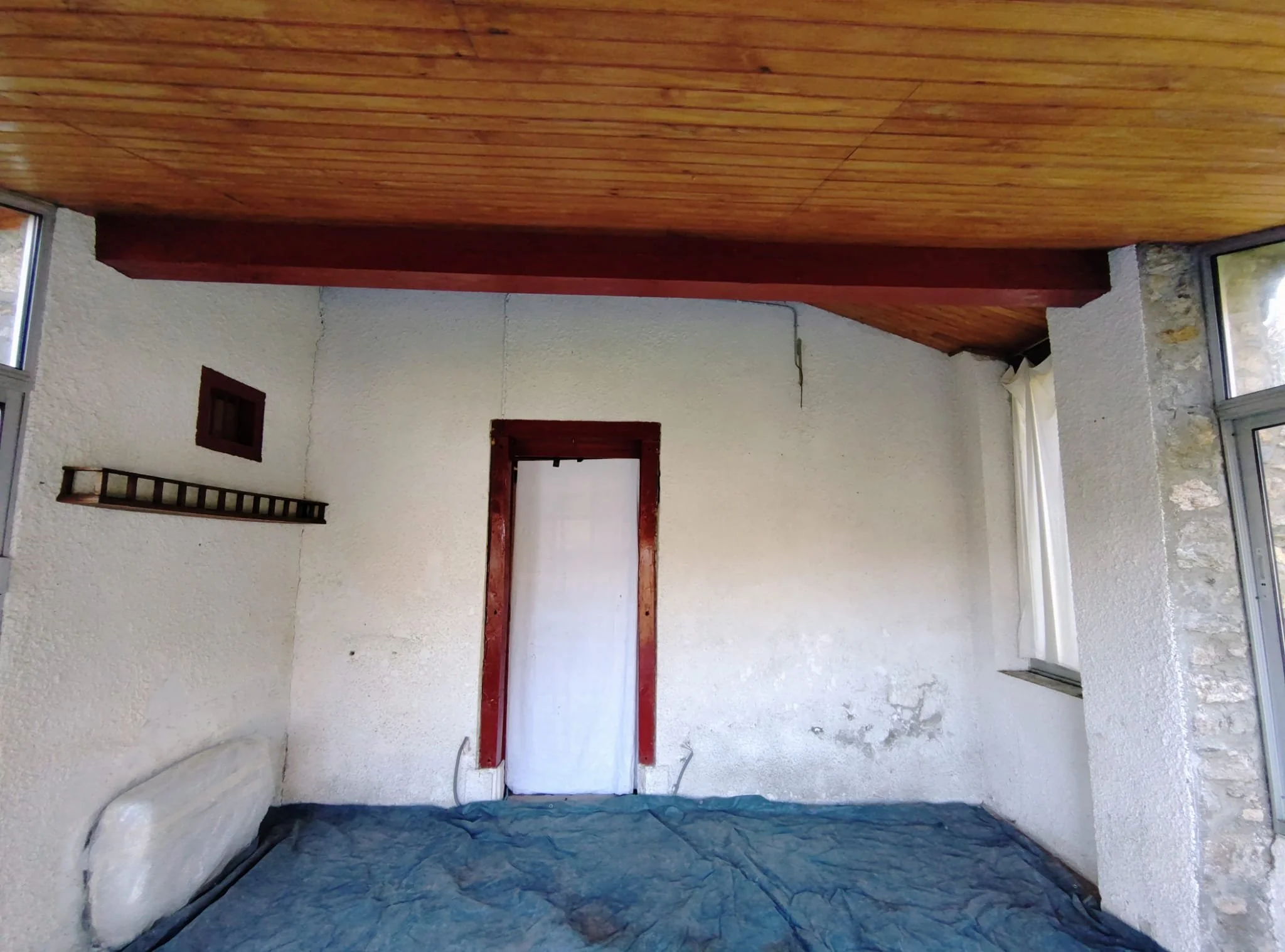 Intérieur d'une pièce vide avec murs blancs, sol recouvert d'une bâche, une fenêtre à droite, une porte au fond avec un cadre en bois, et un étagère en bois sur le mur de gauche.
