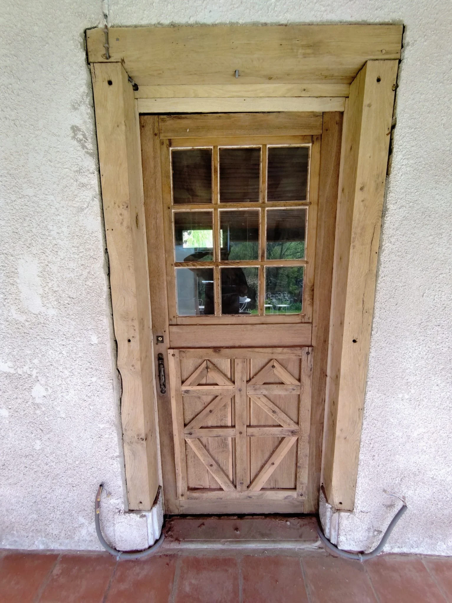 Une porte en bois non peinte avec un panneau en verre divisé en neuf carreaux, entourée d'un cadre en bois. Le mur autour est en plâtre blanc. Deux câbles électriques sortent du sol de part et d'autre de la porte, en bas.