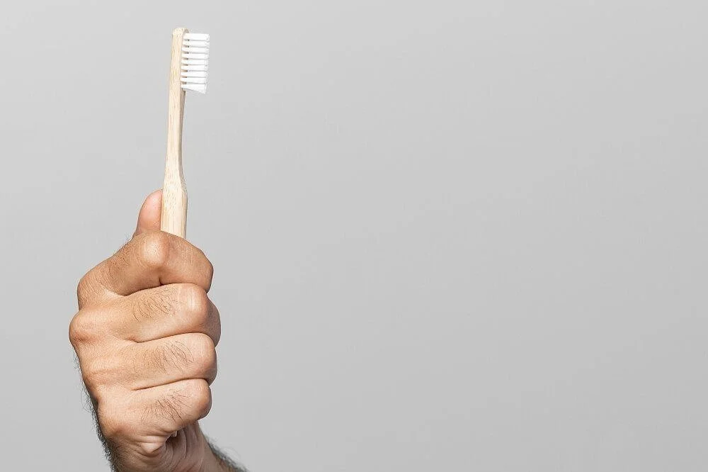 A hand holding a bamboo toothbrush against a gray background.