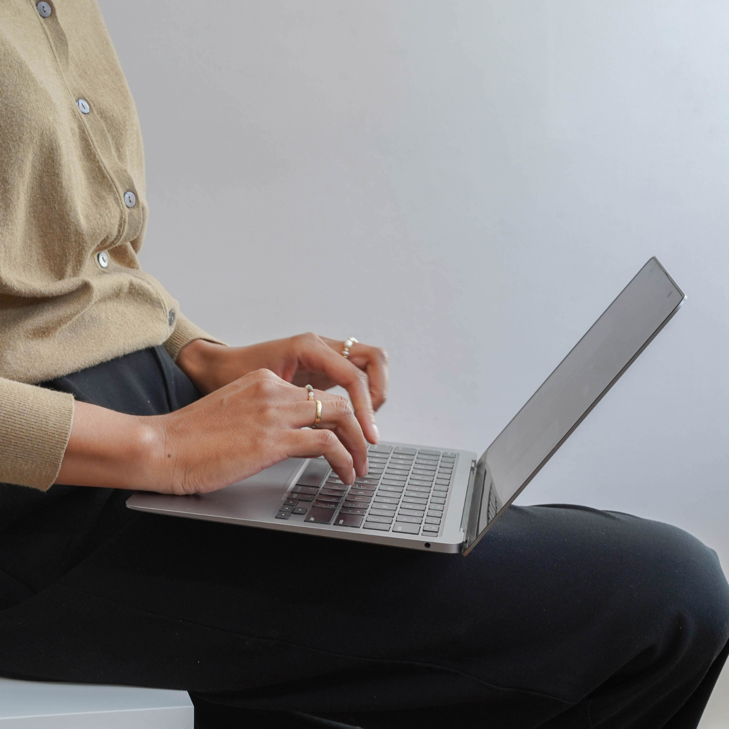 Person wearing a beige sweater working on a silver laptop, sitting against a plain background.