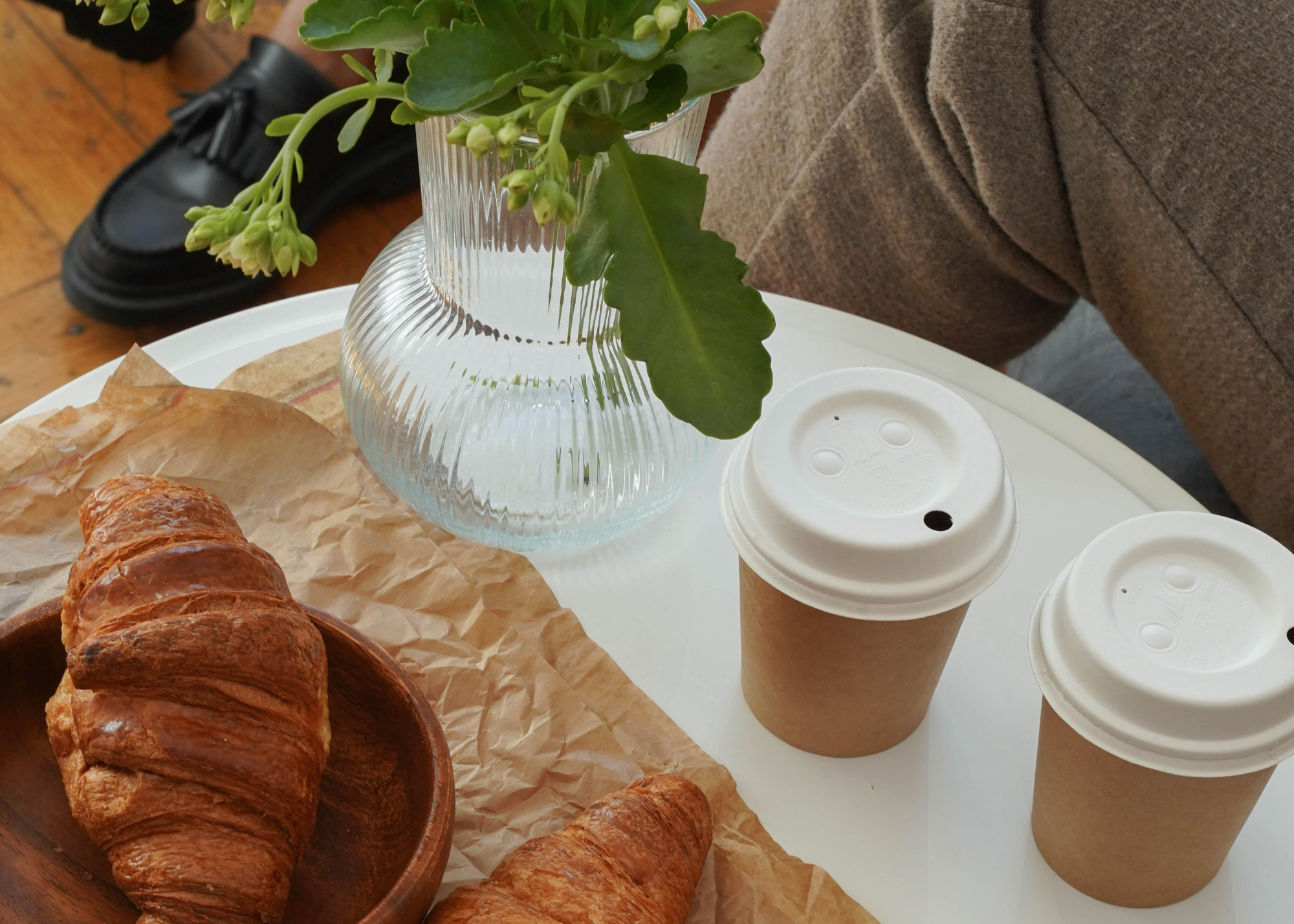 Two disposable coffee cups and a croissant on a white table, with a vase of green leaves, a person in a brown sweater, and a black shoe in the background.