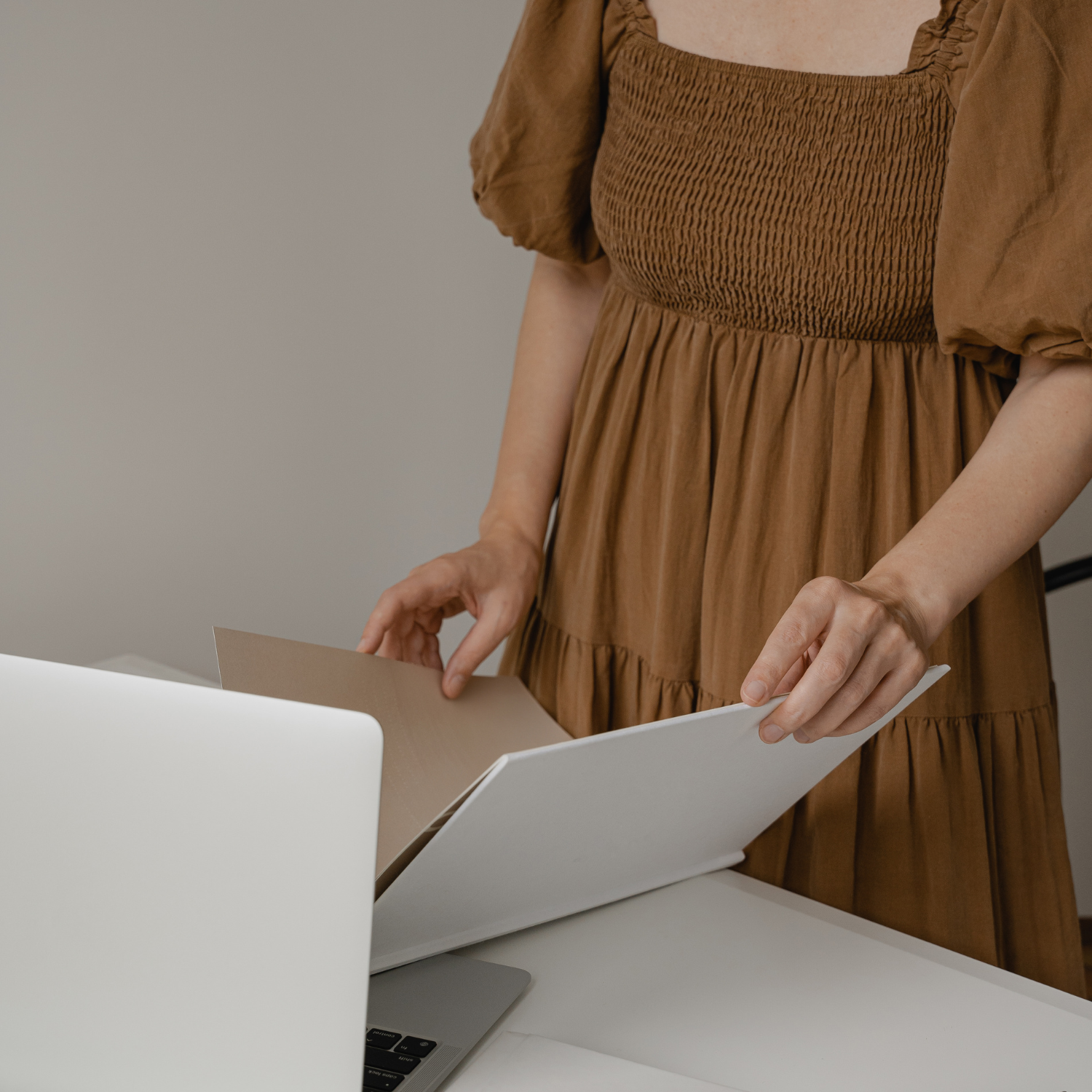 Woman in brown dress flipping through a white folder on a desk with a laptop.