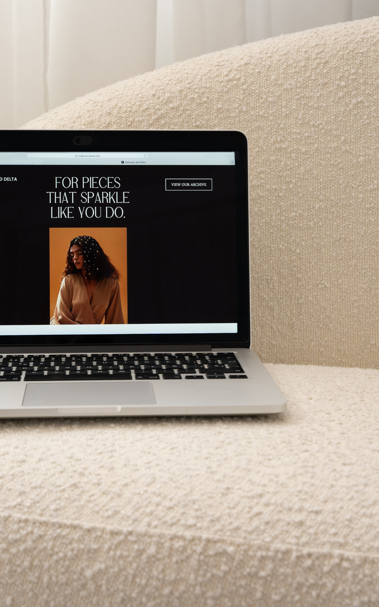 A laptop displaying a website with a woman on the screen, positioned on a textured beige surface, with a light-colored curtain in the background.