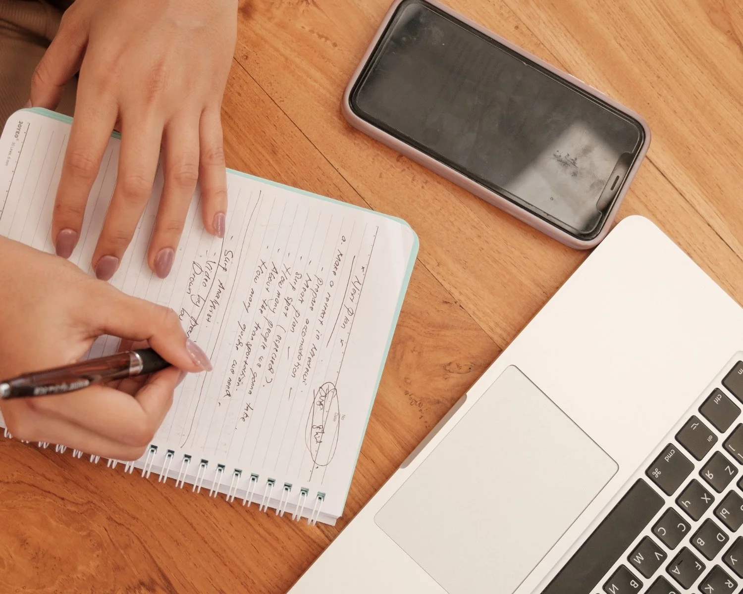 Person writing notes in a spiral notebook on a wooden desk with a laptop, smartphone, and some papers nearby.