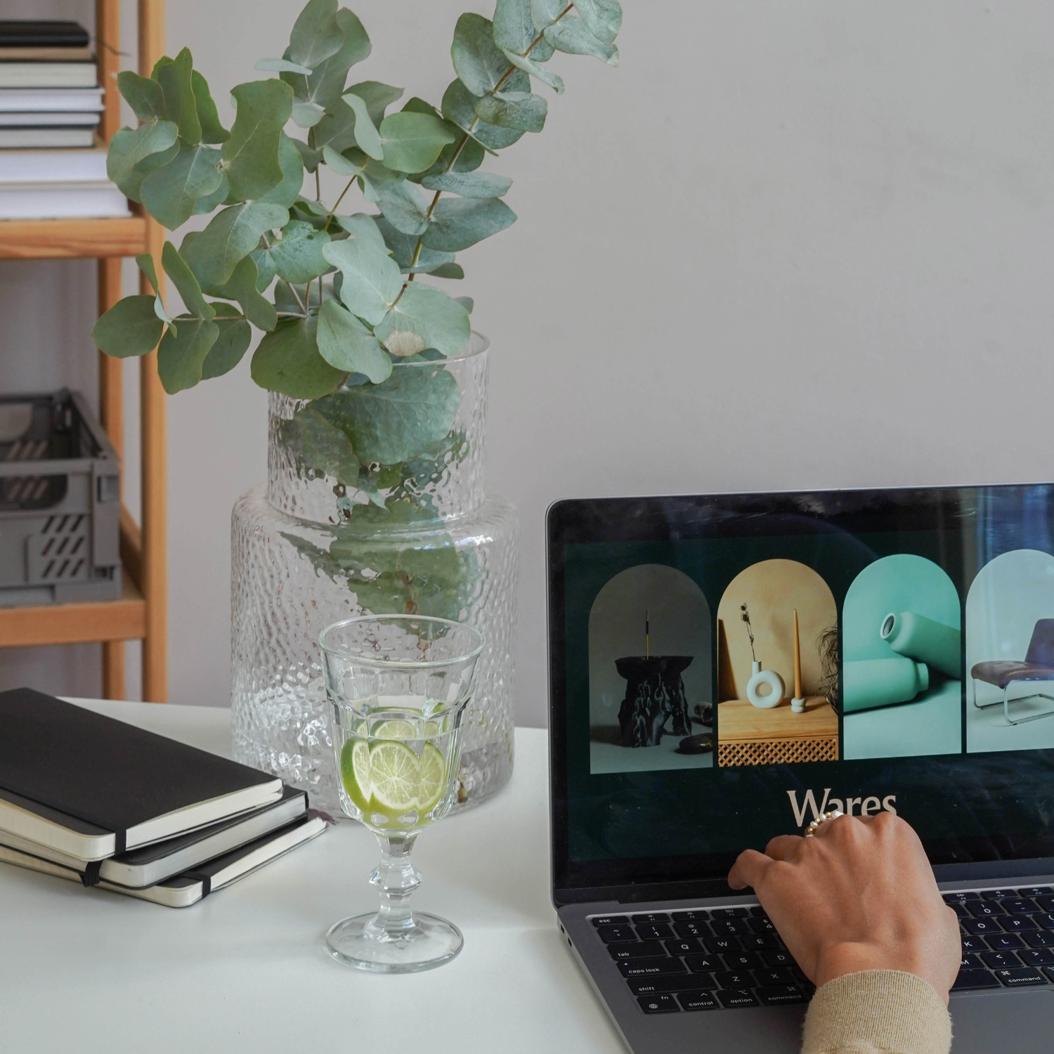 A laptop on a white desk displaying the word 'Wares' and a website with images of modern home decor in a grid layout. Next to the laptop is a glass of water with lemon slices. A large glass vase with eucalyptus leaves and a small wooden shelf with stacked books are in the background.