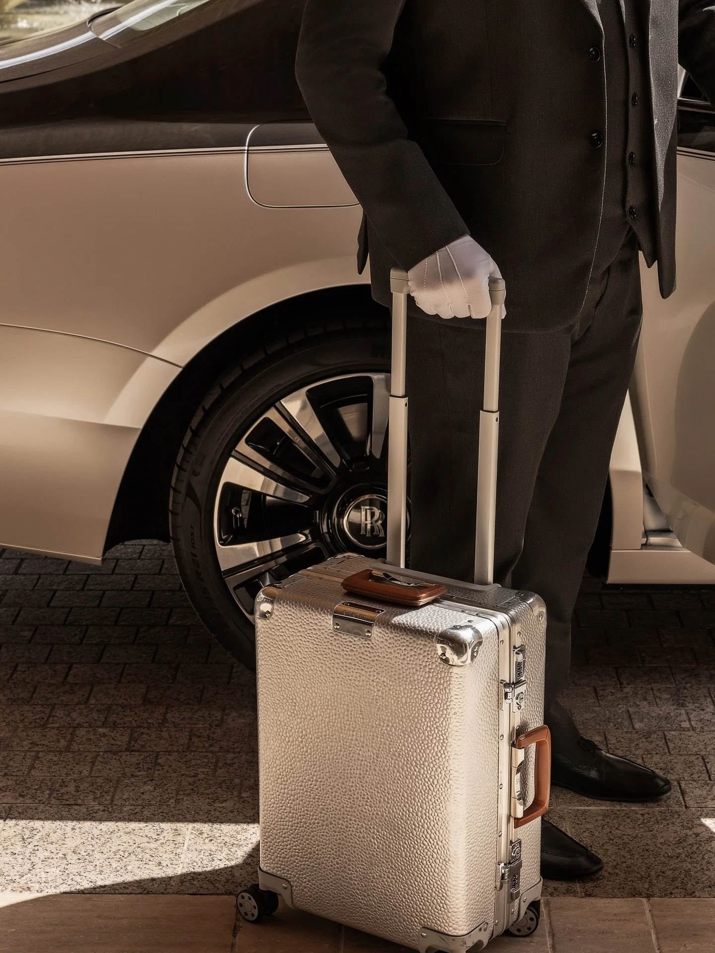 A man in a black suit carrying a suitcase in front of a luxury car.