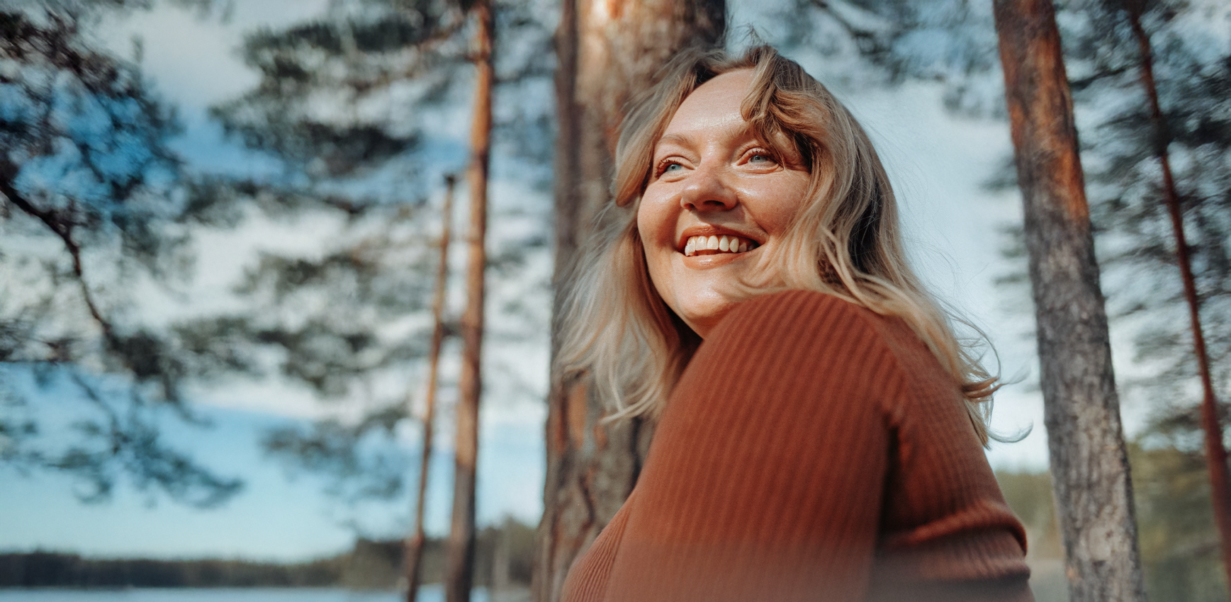 Ihminen hymyilee ulkona, taustalla puita ja sininen taivas.