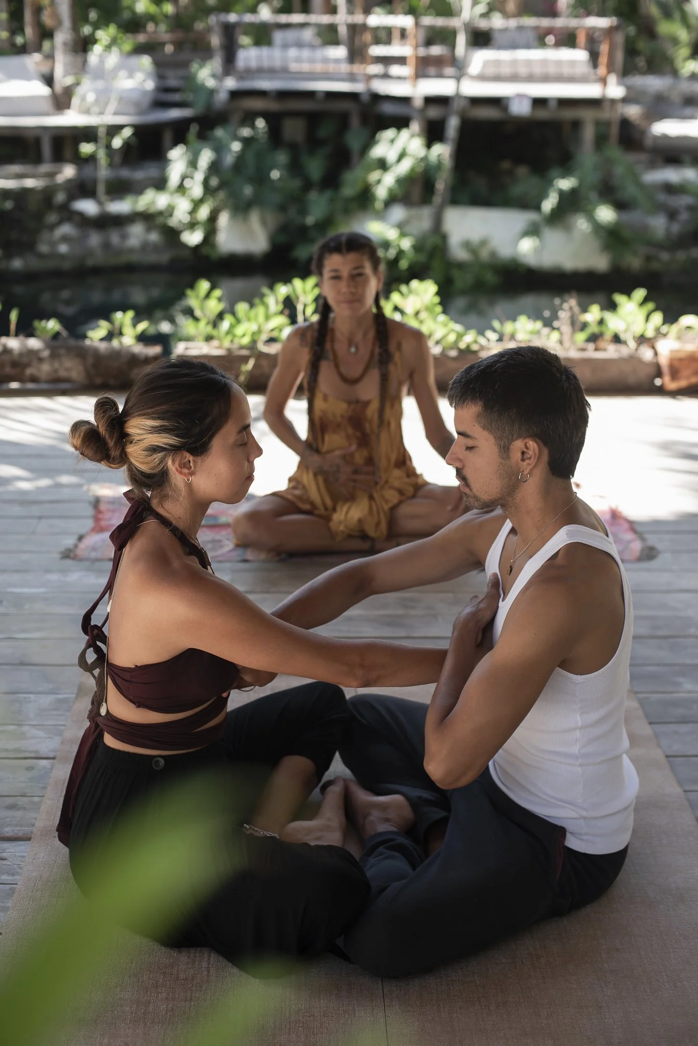 Two people sitting cross-legged on a wooden platform, facing each other with their eyes closed, practicing psychedelic breathwork specifically for couples, with a medicine woman leading the session.