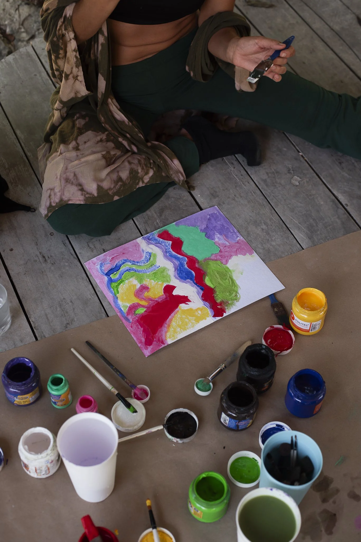 Person sitting on a wooden floor painting a colorful abstract artwork with various paint bottles, brushes, and supplies around them.