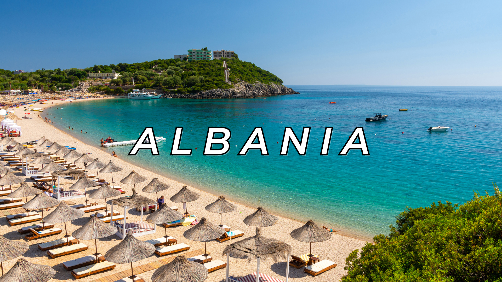 A scenic beach in Albania with clear turquoise water, straw umbrellas, lounge chairs, and a hillside with buildings and greenery in the background.