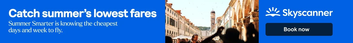 Banner ad for Skyscanner featuring a city street with historical buildings, a clock tower, and a crowd of people, promoting low fares for summer travel.
