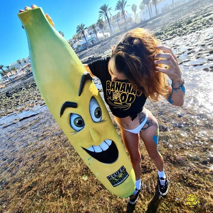 Mujer en la playa sosteniendo un inflable en forma de plátano con cara sonriente, vistiendo una camiseta negra con letras amarillas que dicen 'Banana Mood', gafas de sol en la cabeza y pantalones cortos blancos.