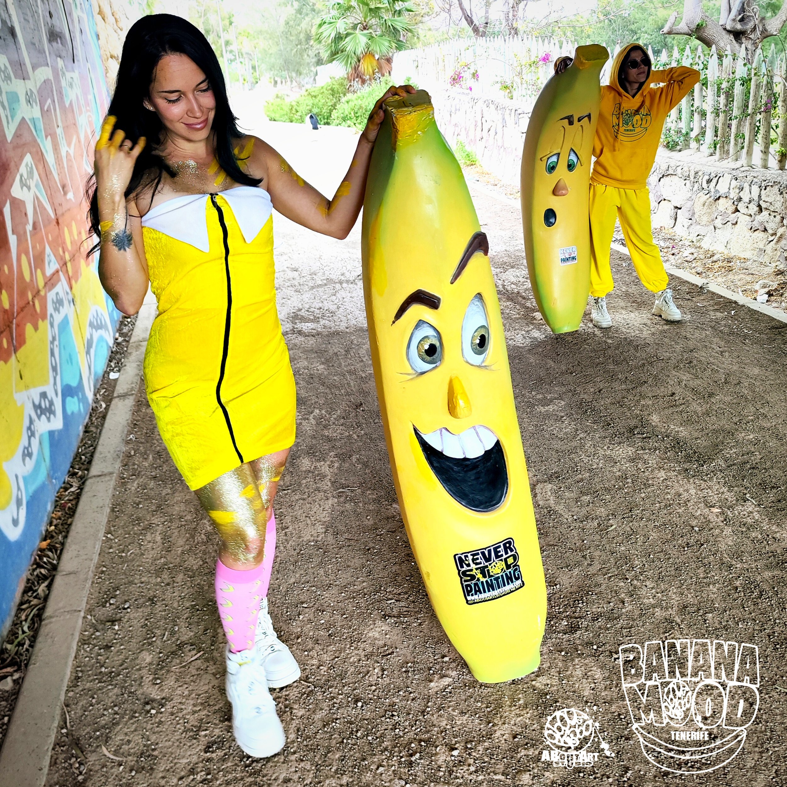 Dos personas disfrazadas con ropa amarilla, una mujer y un hombre, sosteniendo esculturas de plátanos con caras felices en un entorno al aire libre con muro pintado y plantas.
