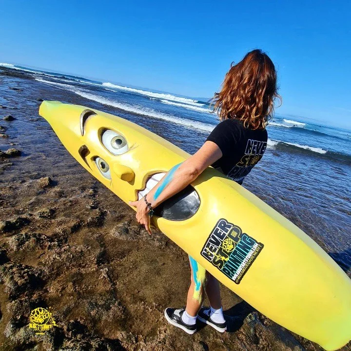 Persona con cabello rizado de color castaño claro sosteniendo una tabla de surf amarilla con dibujos y efectos faciales en la playa