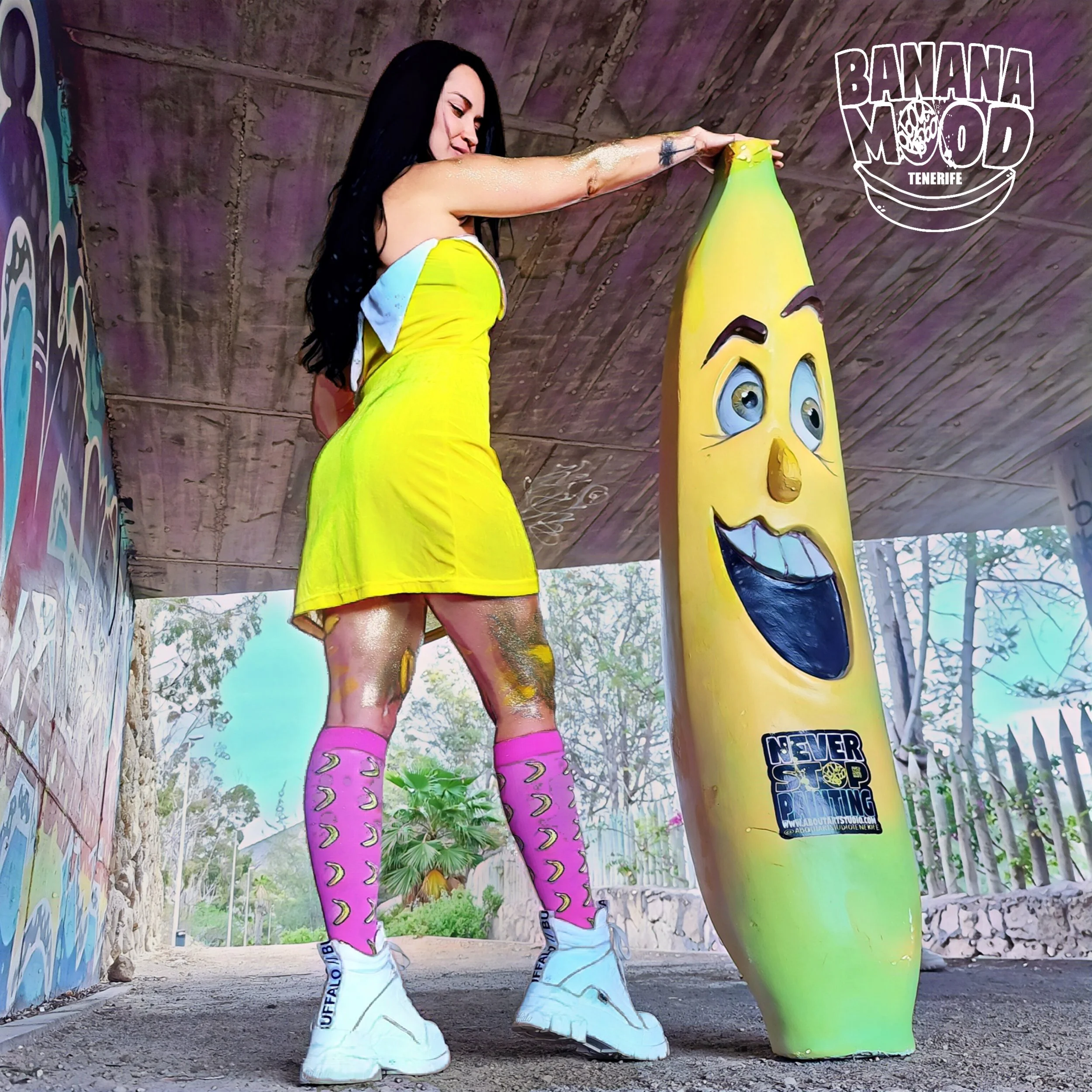 Mujer con vestido amarillo y medias con patos rosas, sosteniendo una figura de zanahoria con rostro sonriente bajo un puente con graffiti