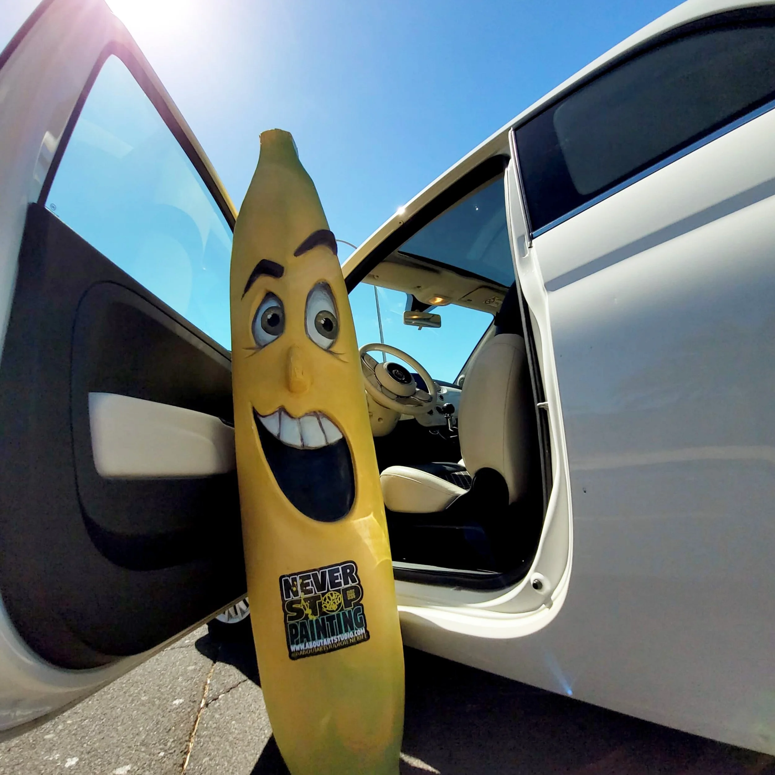 Banana con expresión facial sonriente y mensaje publicitario, junto a un coche con puertas abiertas bajo un cielo azul y sol brillante.
