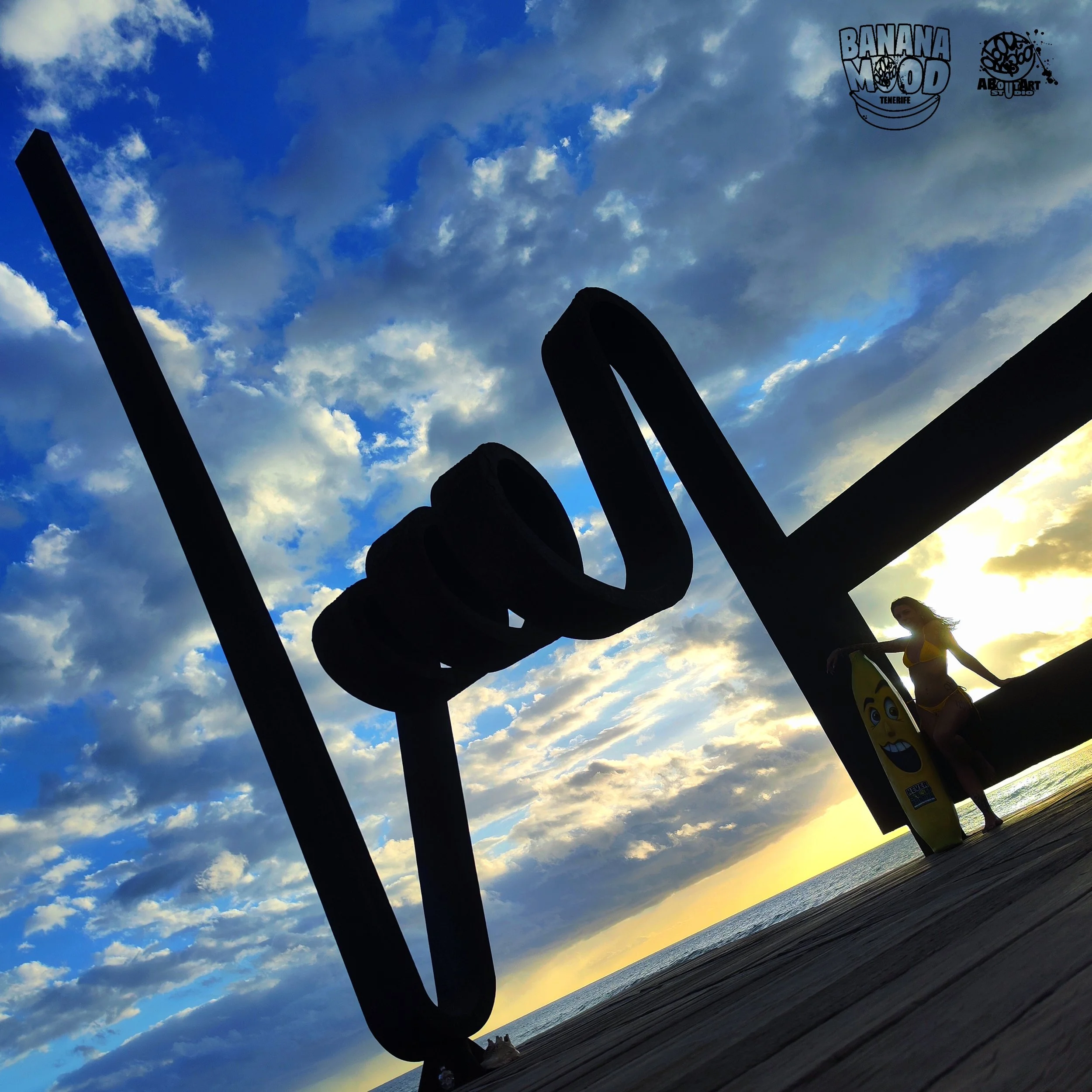 Silhouette de letras volcadas que dicen 'LOVE' en un muelle frente al mar durante el atardecer, con un cartel de surfing y una mujer en Bikini, en un día con cielo parcialmente nublado.