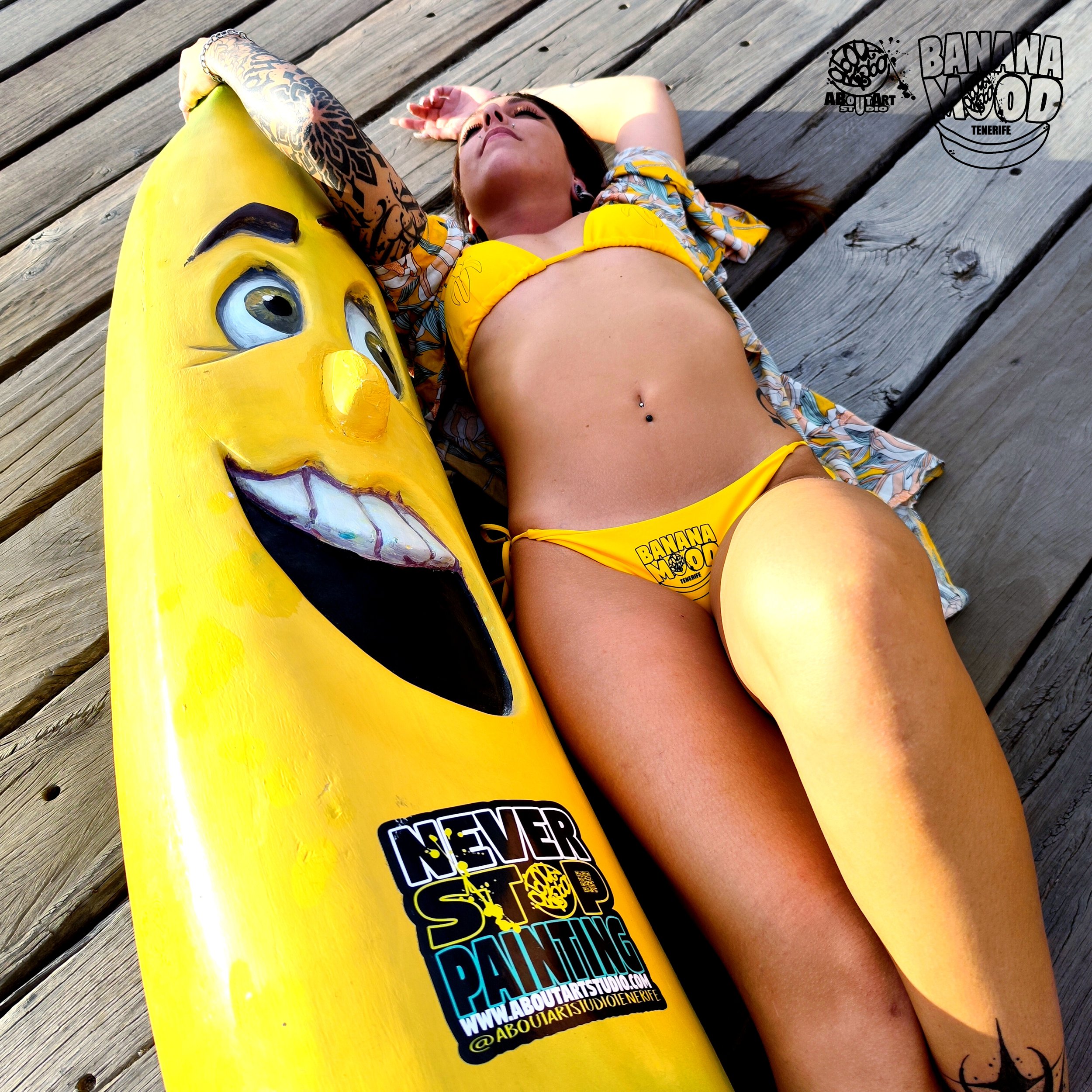 Joven mujer con bikini amarillo, tumbada en madera, descansando junto a una tabla de surf con cara sonriente y texto de 'Never Stop Painting', en Tenerife.