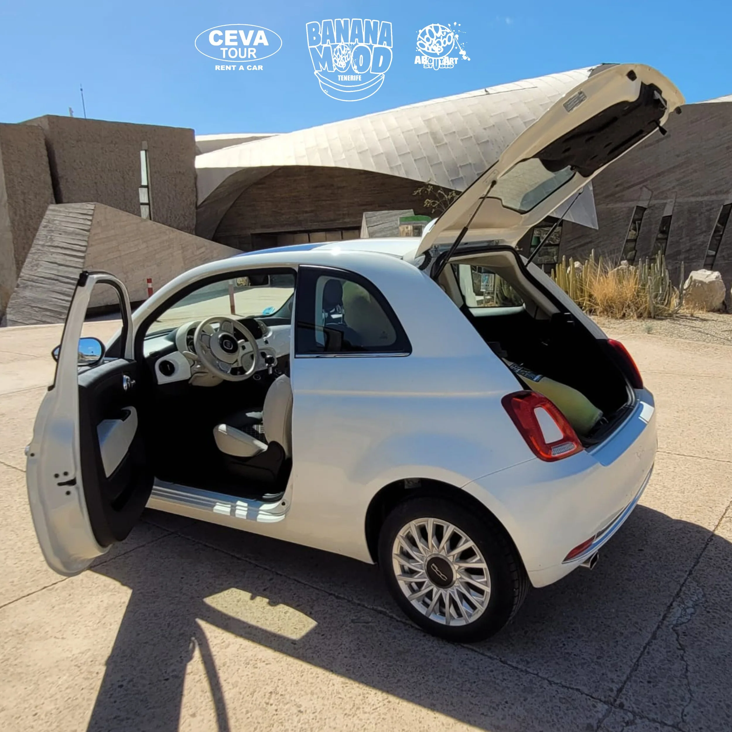 Coche pequeño blanco con la puerta y el maletero abiertos estacionado en un área pavimentada, con un edificio de arquitectura moderna en el fondo y algunos cactus y plantas.