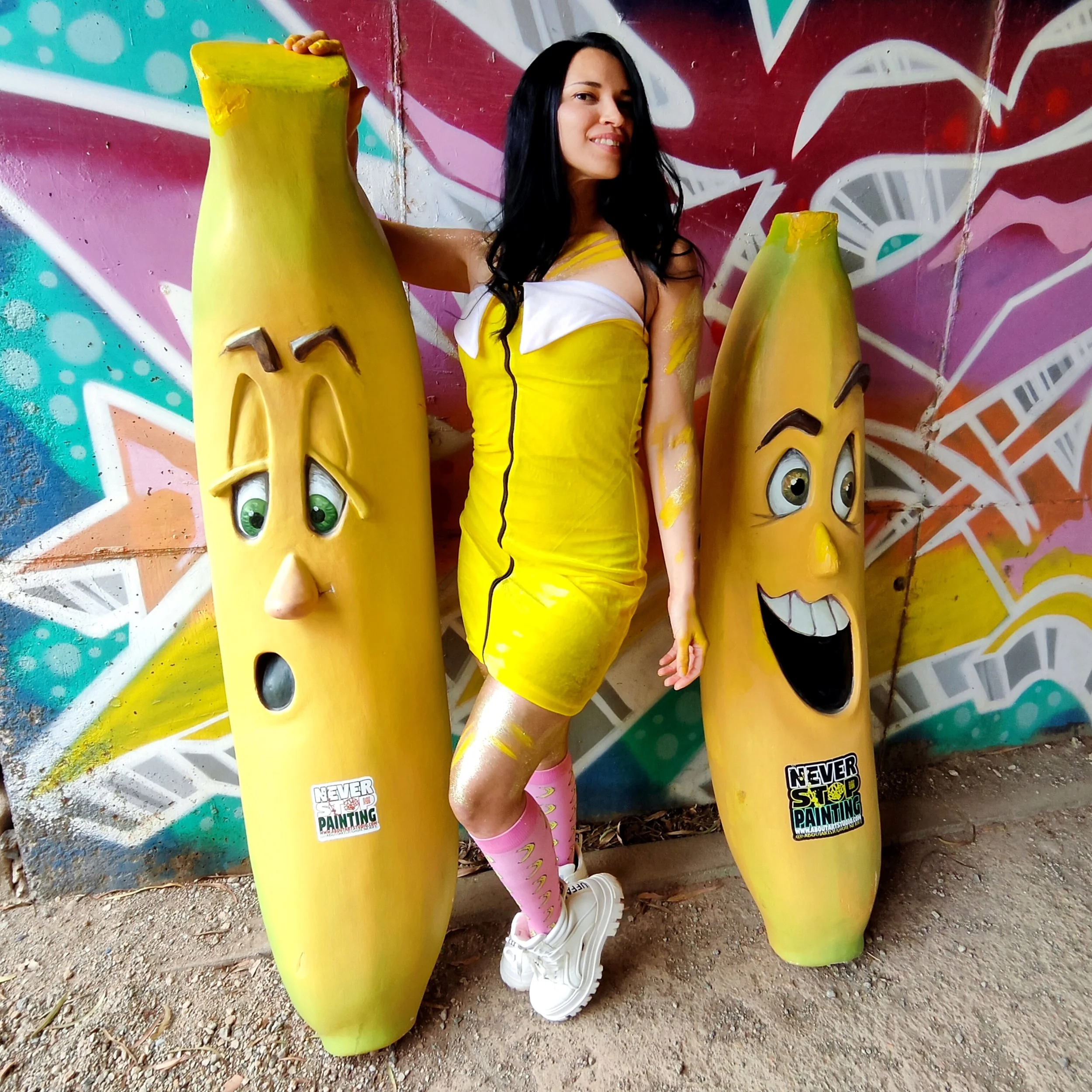 Mujer con ropa amarilla y rosa, con dos esculturas de plátano con caras haciendo expresiones diferentes, en un fondo de grafiti colorido.