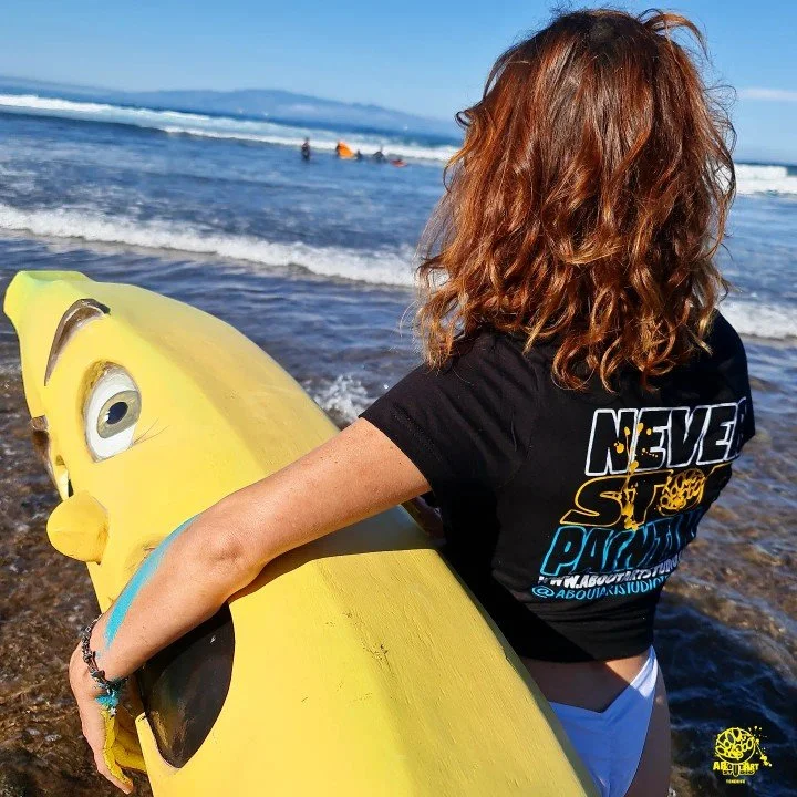 Mujer con cabello rizado color castaño oscuro sosteniendo una tabla de surf en la orilla del mar, vista de espaldas, con el océano y el cielo azul en el fondo.