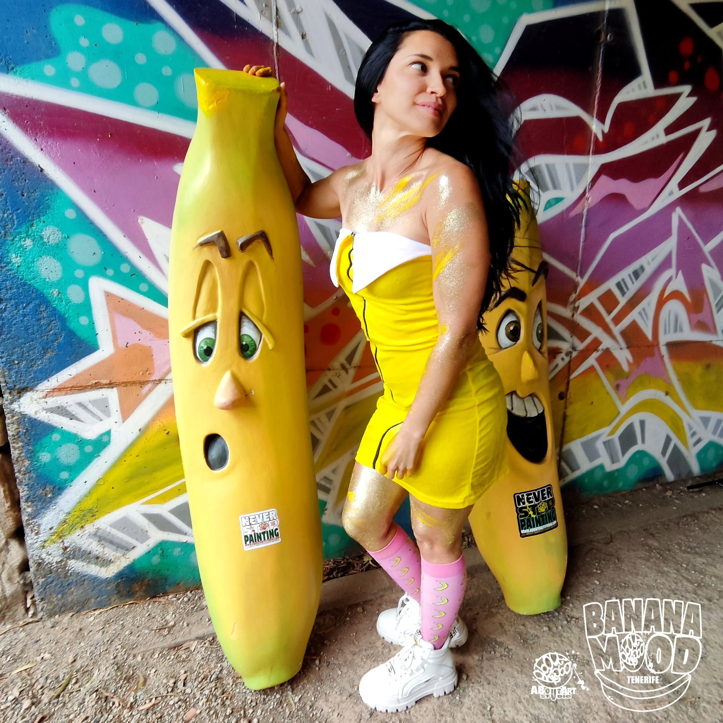 Una mujer con vestido amarillo y medias rosas posa junto a esculturas de plátanos con caras expresivas y un fondo de graffiti colorido.