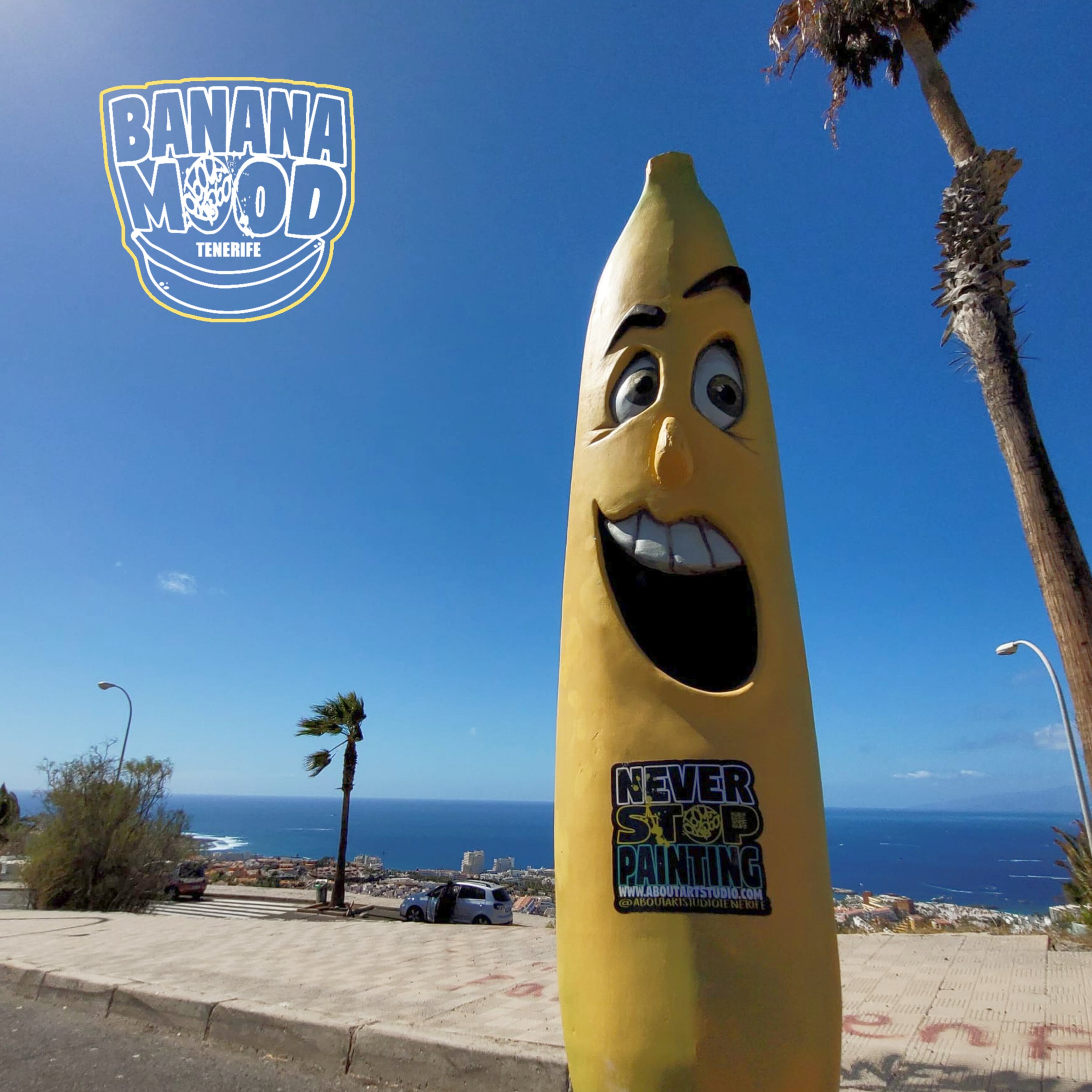 Escultura de una banana amarilla con cara sonriente, ojos grandes y cejas negras, en un mirador con vista al mar y a la ciudad, con un logo que dice 'Banana Mood Tenerife' y una pegatina que dice 'Never Stop Painting'.