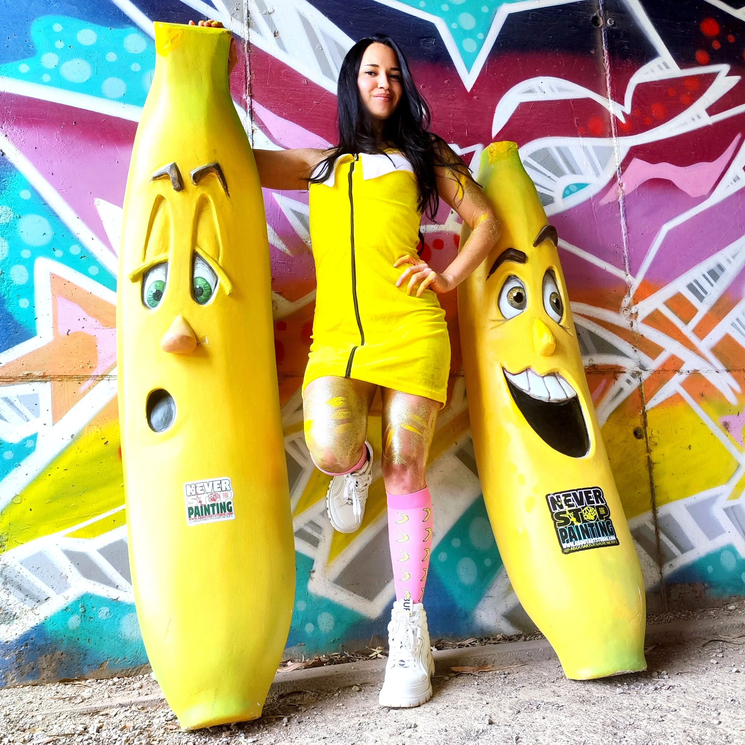 Mujer vestida de amarillo, posando entre dos postes de jabón con caras caricaturescas, en un mural colorido de fondo.