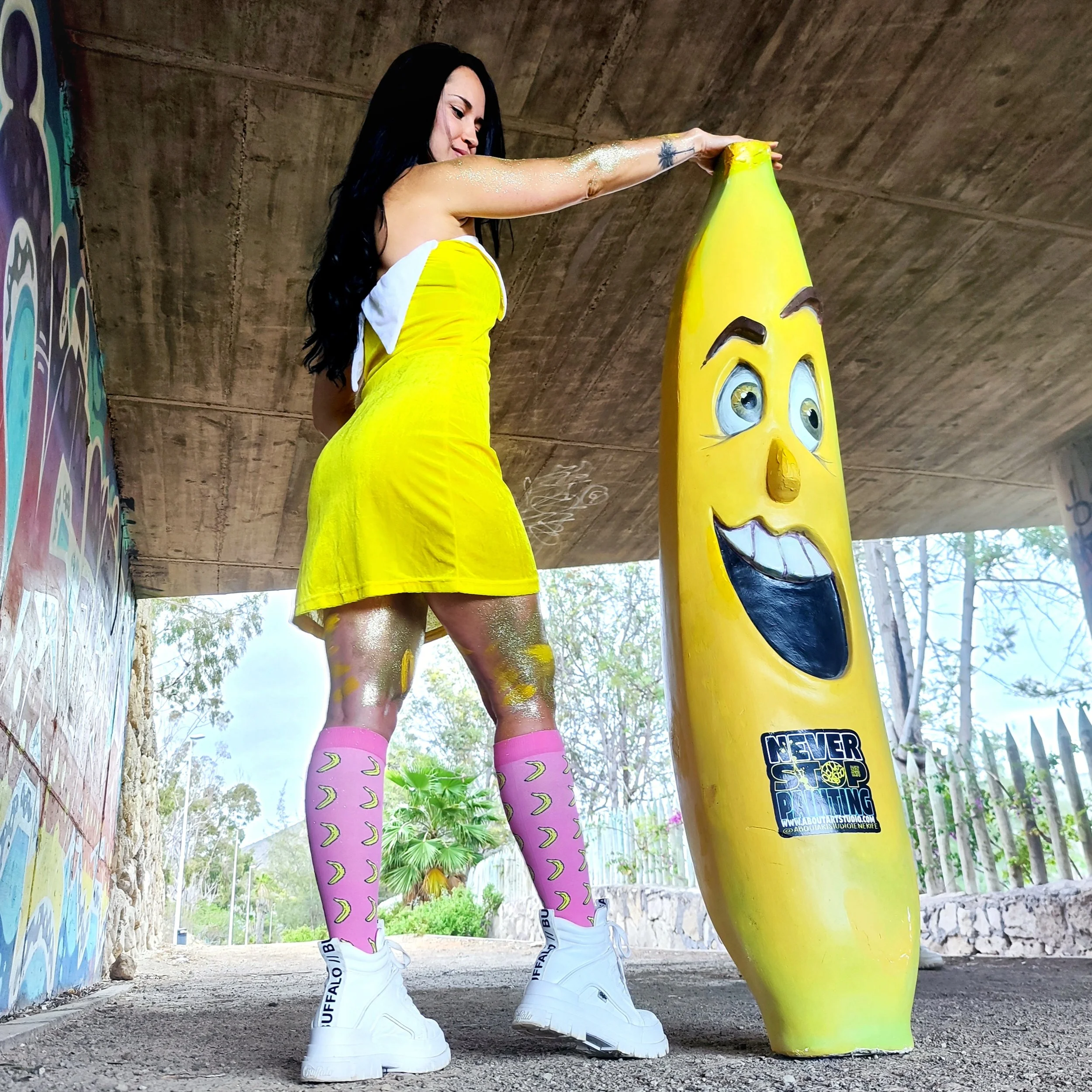 Una mujer con vestido amarillo y calcetas rosadas con plátanos, usando botas blancas, interactúa con una escultura de una banana con cara sonriente, en un lugar con grafitis y árboles al fondo.
