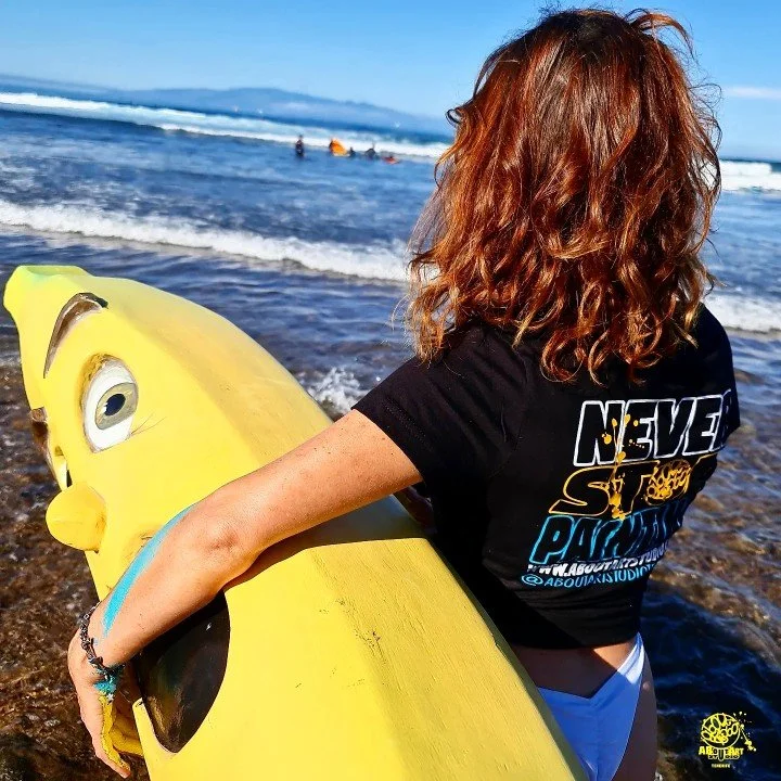 Persona con cabello rizado de color castaño claro, de espaldas, sosteniendo una tabla de surf amarilla con diseño de Bob Esponja en la orilla del mar