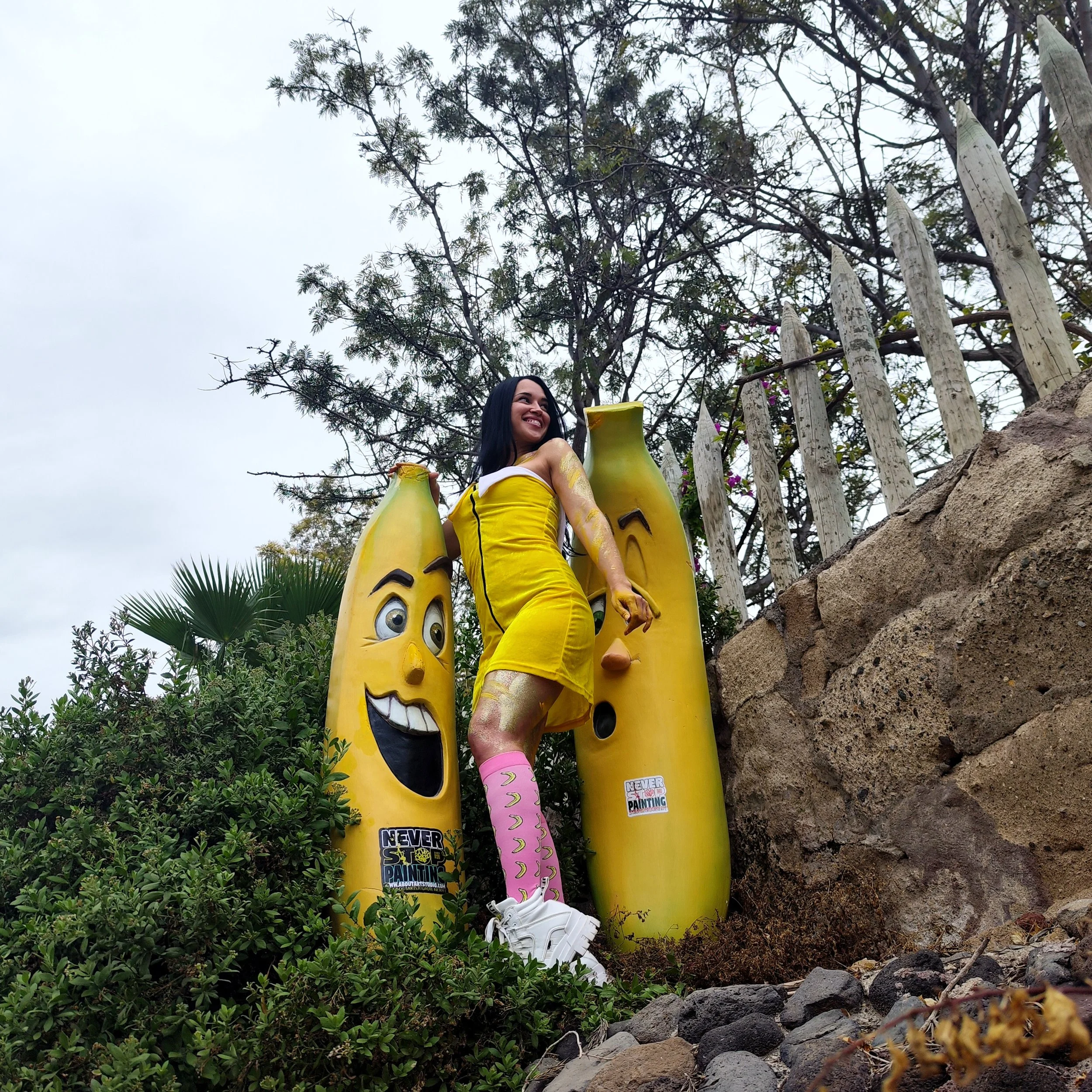 Mujer disfrazada de banana amarilla posando frente a decoraciones de plátanos con caras expresivas, en un ambiente exterior con árboles y una cerca de madera.