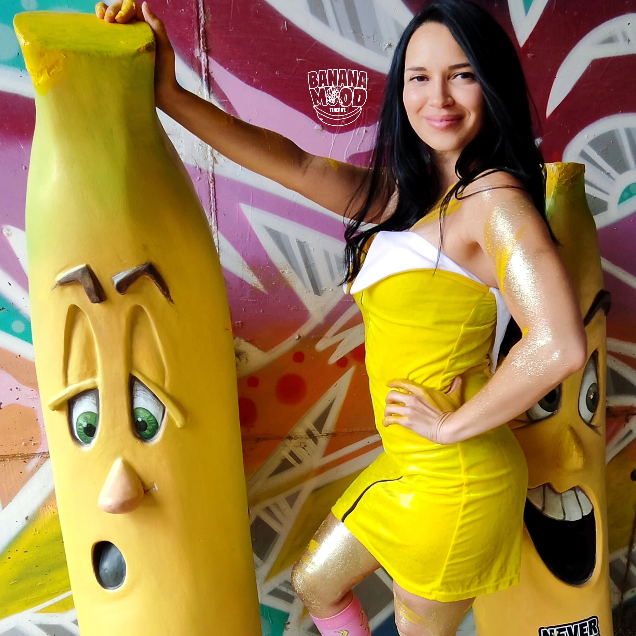 Una mujer con vestido y accesorios amarillos con detalles dorados, posando junto a esculturas de plátanos con expresiones faciales, en un fondo con graffiti colorido.