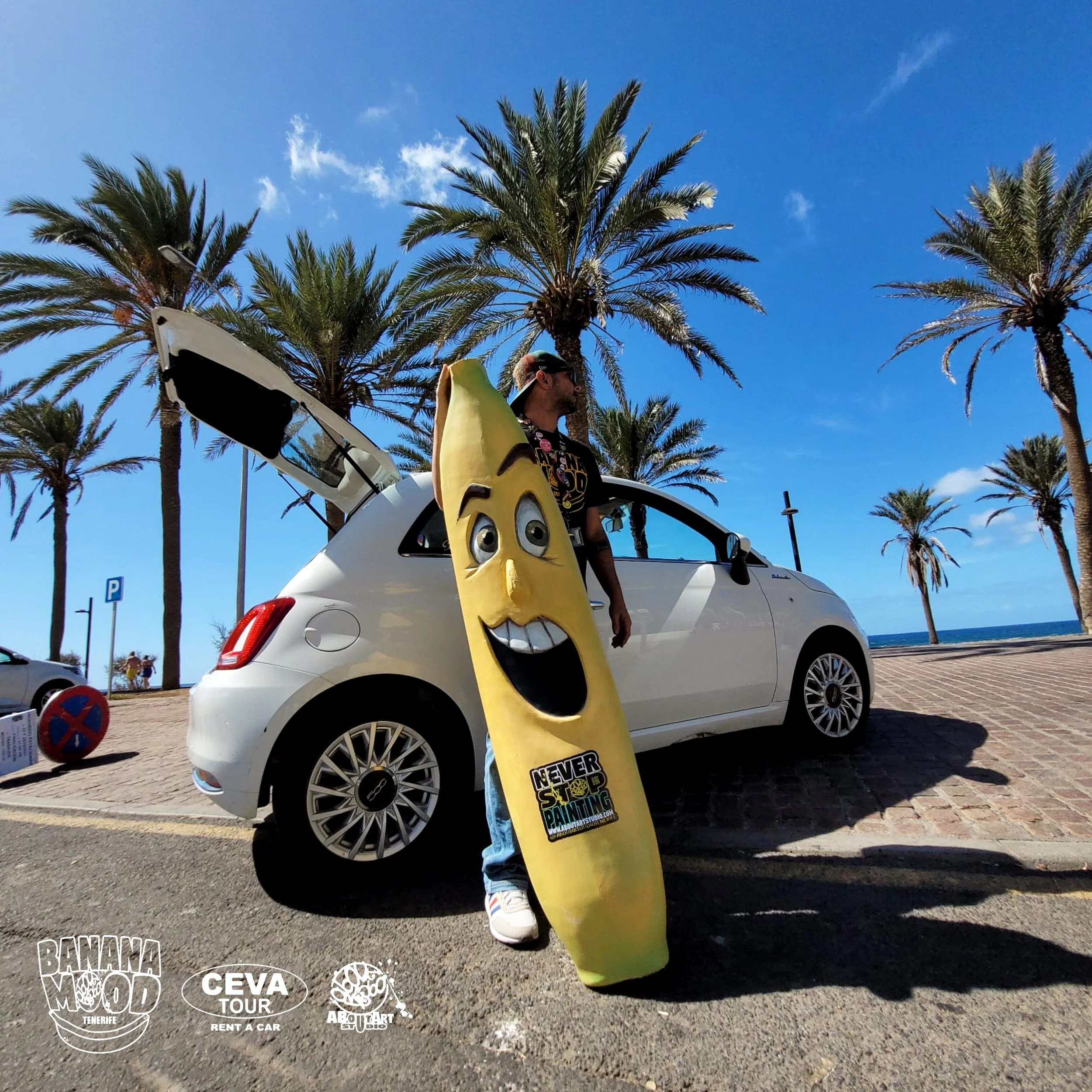 Hombre con gafas de sol y camiseta estampada, sosteniendo una tabla de surf con rostro sonriente, de color amarillo, en una zona costera con palmeras y un cielo azul, junto a un coche blanco con la puerta trasera abierta.