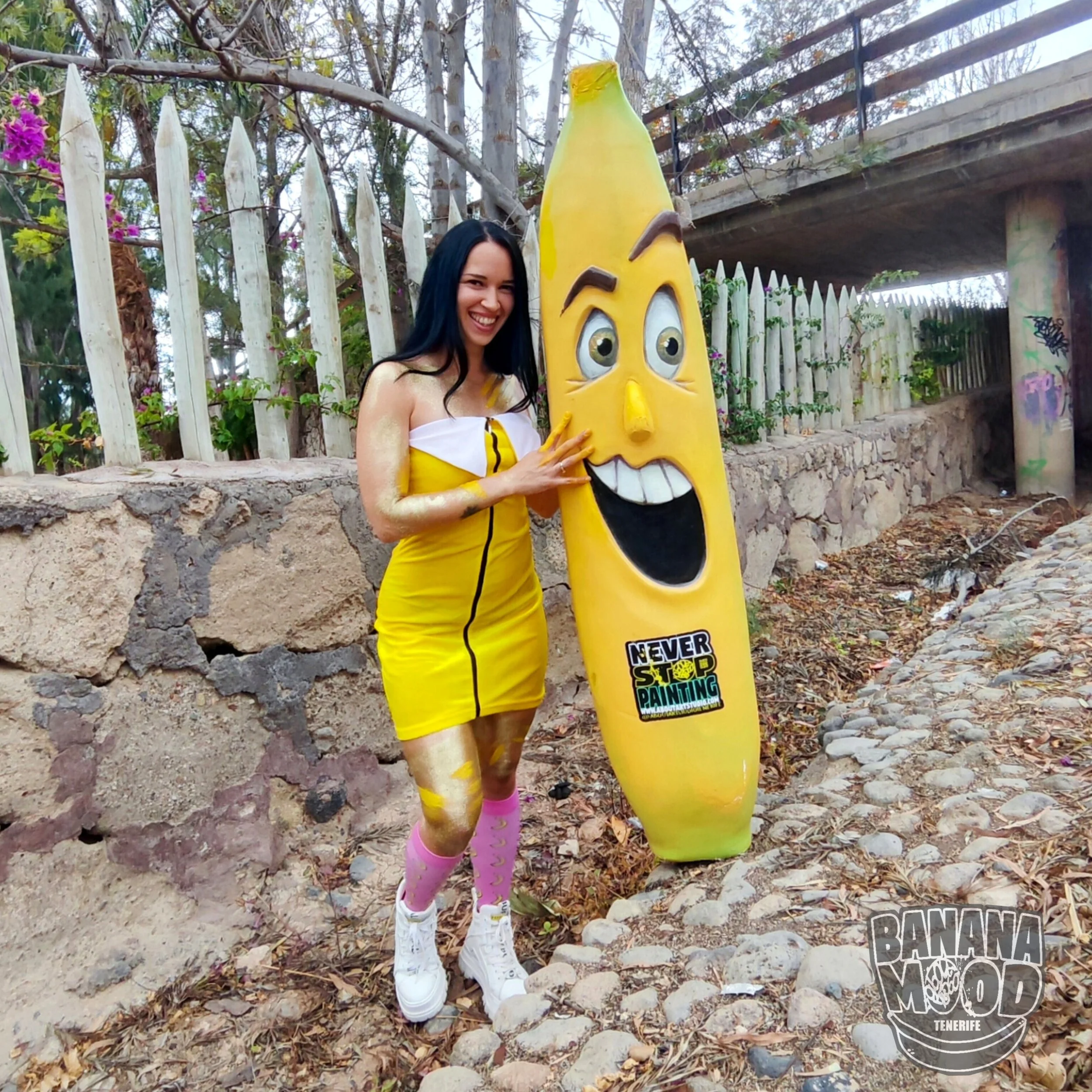 Mujer disfrazada de plátano posando con una estatua de plátano con expresión feliz al aire libre en Tenerife, con fondo de valla de madera y vegetación.