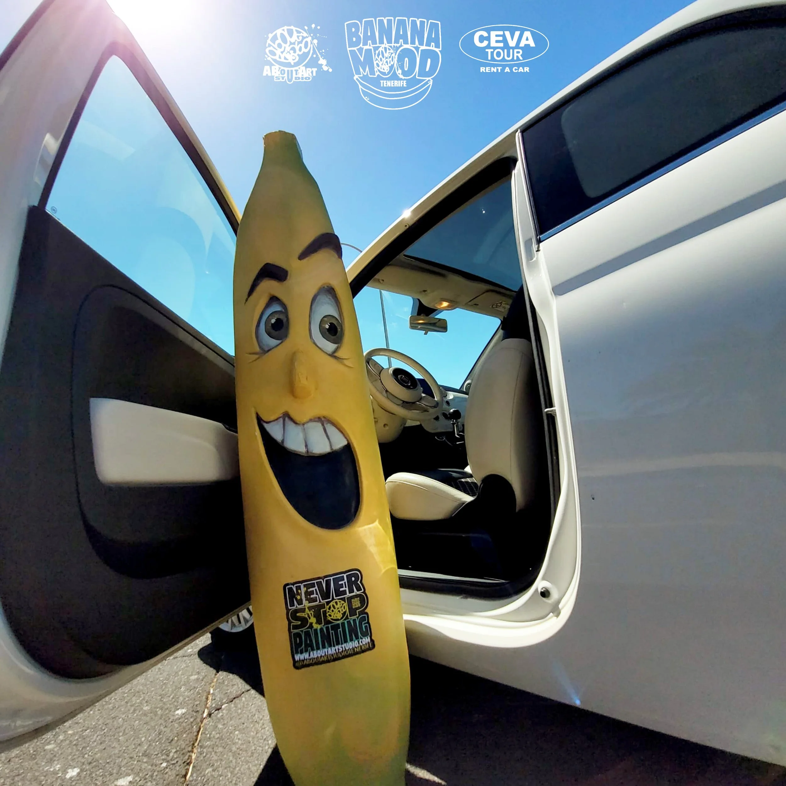 Una banana con rostro antropomórfico y expresión alegre, apoyada contra la puerta de un coche blanco con las puertas abiertas, en un día soleado. En el cielo se ven varios logotipos y textos relacionados con turismo y arte, como 'Banana Mood Tenerife