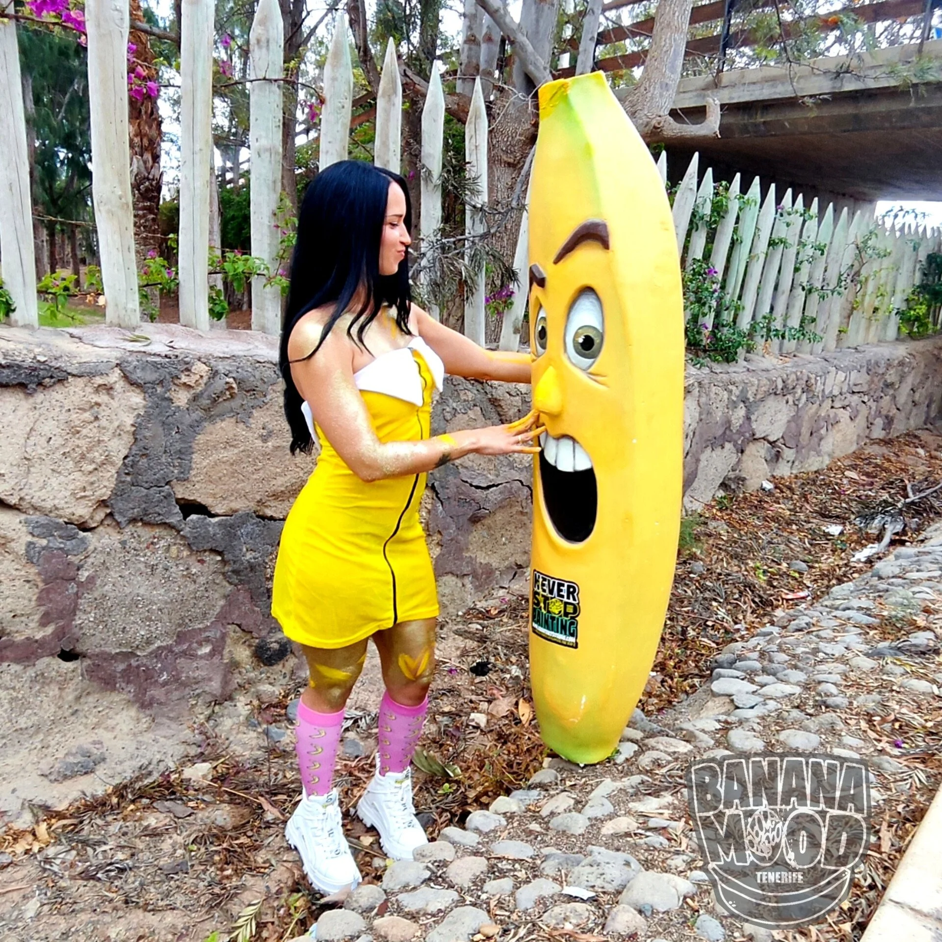 Una mujer con un vestido amarillo y calcetas rosas interactuando con una figura de arte callejero que representa una banana con una cara y boca grande en una zona exterior con piedra y vegetación.