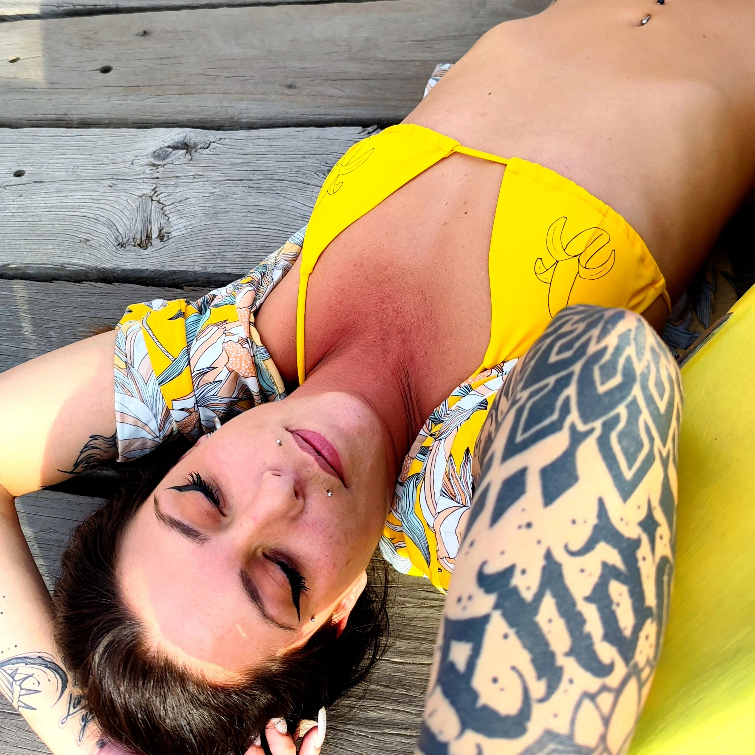 Mujer con tatuajes y piercings descansando en una madera, vistiendo bikini amarillo y falda con estampado floral, con el cabello oscuro y levantando un brazo.