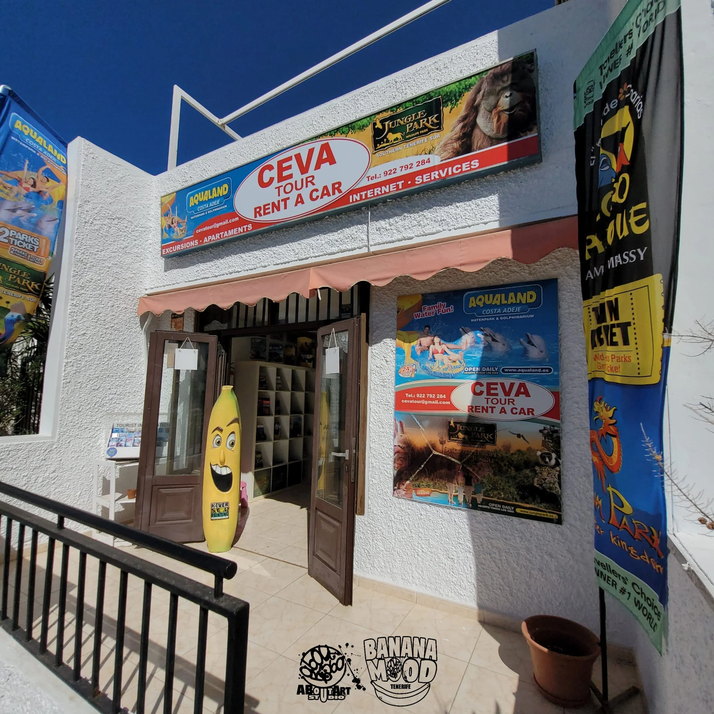 Fachada de una tienda con señalización que anuncia alquiler de autos y tours en Ceuta, con cartelera promocional de Aqualand en Costa Adeje, Tenerife, y decoración con una banana con cara sonriente y un banner vertical colorido.